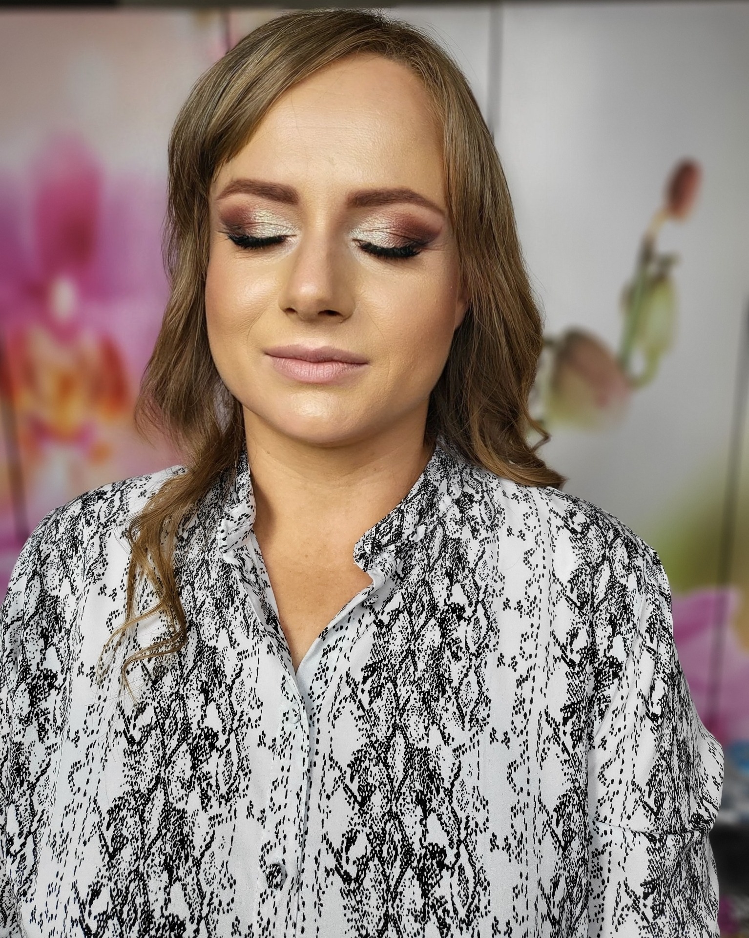 Portret kobiety z zamkniętymi oczami, prezentujący delikatny, błyszczący makijaż oczu w odcieniach brązu i złota, z subtelnym podkreśleniem rzęs i naturalnym makijażem ust, ubrana w koszulę...