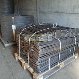 Paleta z uformowanymi prętami zbrojeniowymi w kształcie litery L, zabezpieczona białymi taśmami, obok zwoje drutu zbrojeniowego w magazynie.