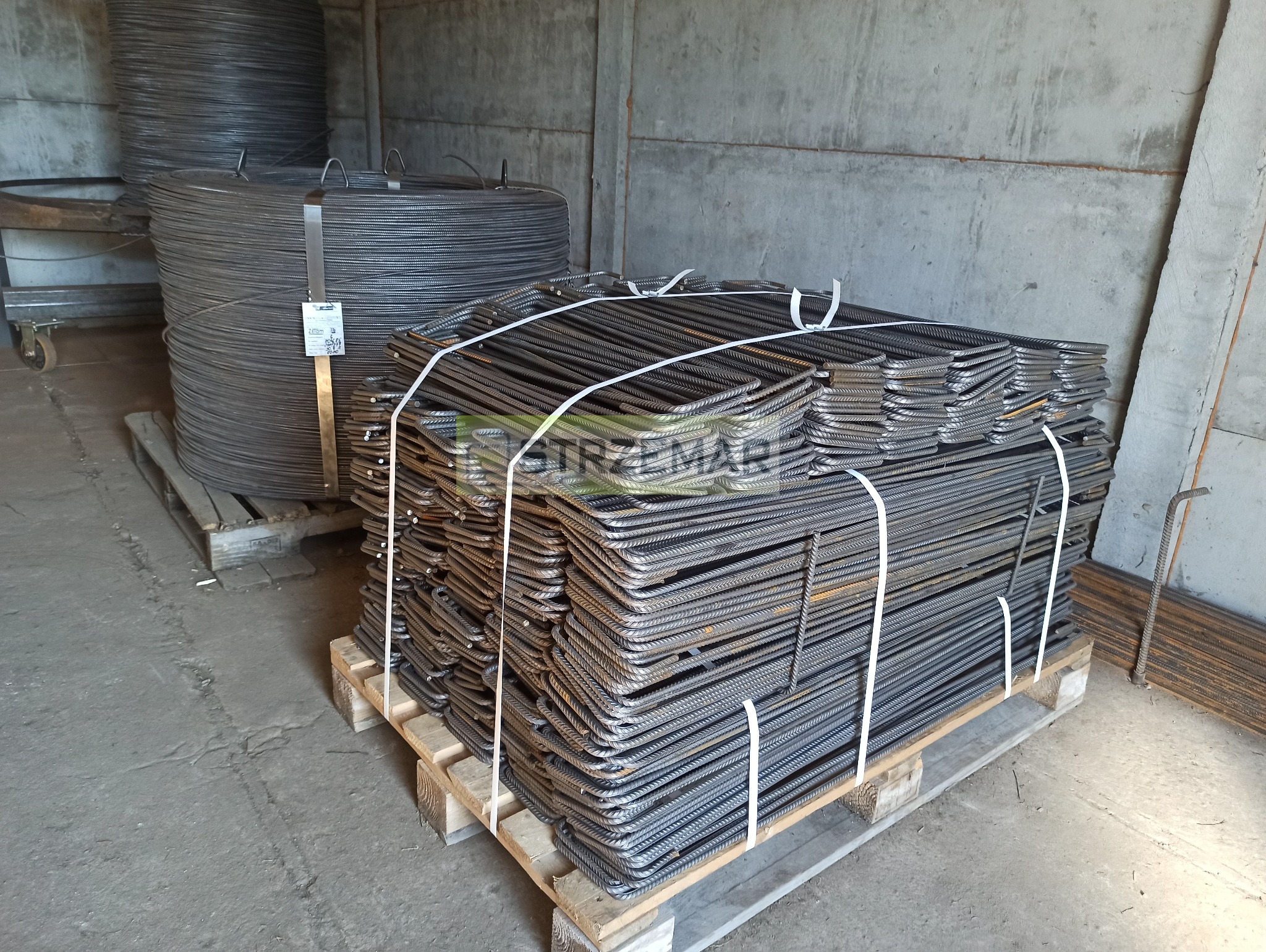 Paleta z uformowanymi prętami zbrojeniowymi w kształcie litery L, zabezpieczona białymi taśmami, obok zwoje drutu zbrojeniowego w magazynie.