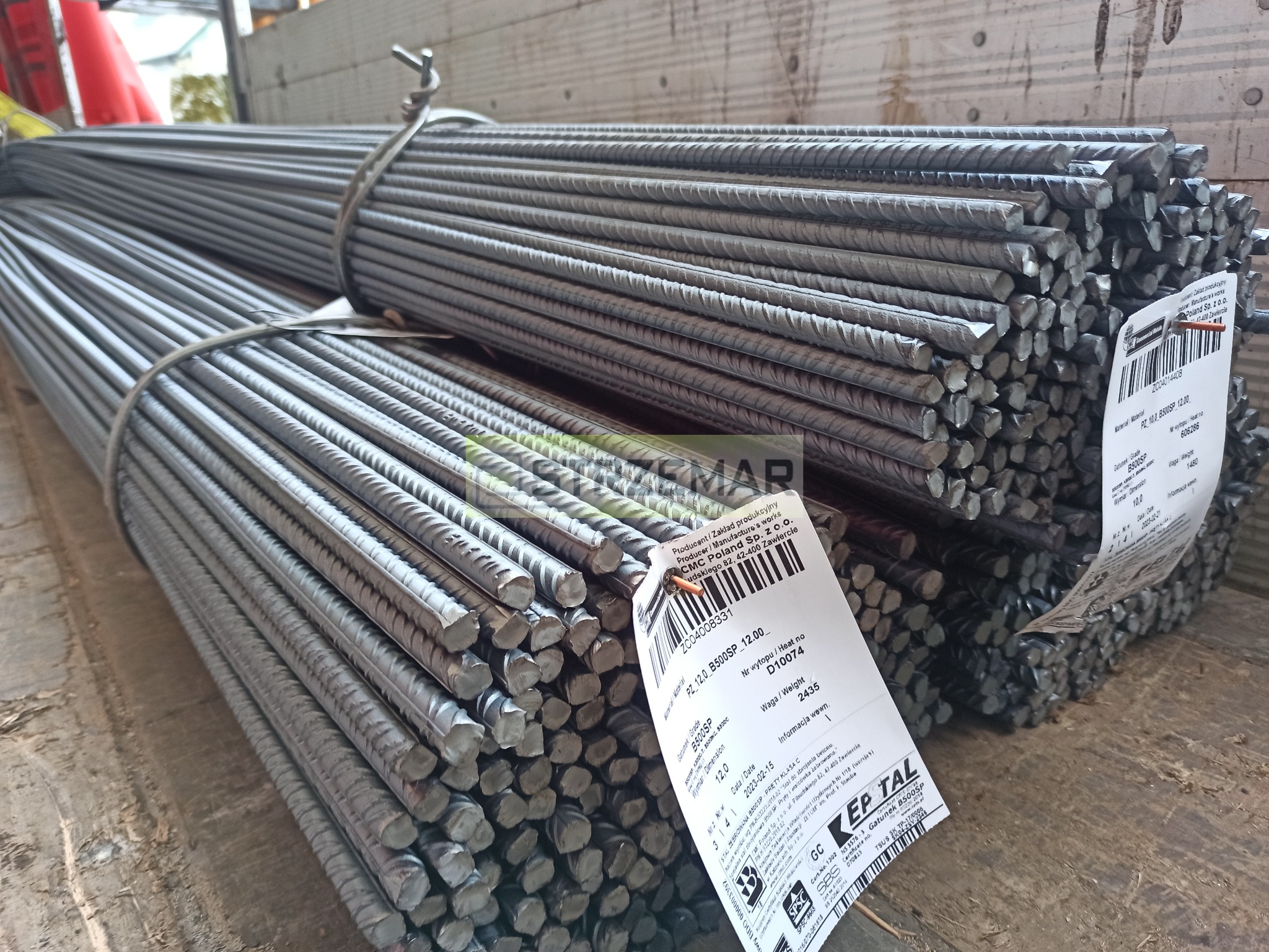 Dwa pakiety żebrowanych prętów stalowych, związane drutem, z etykietami producenta na drewnianej powierzchni ładunkowej. Widoczne informacje o gatunku stali B500SP i wadze.