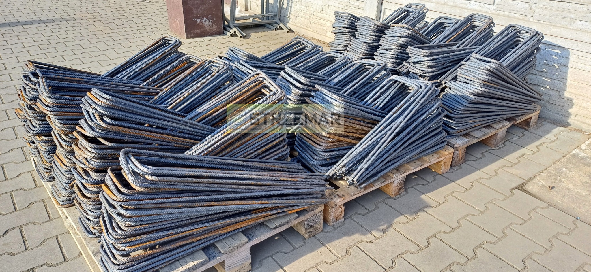 Stosy prefabrykowanych, giętych prętów zbrojeniowych ułożone na drewnianych paletach na kostce brukowej, widoczne ślady rdzy.