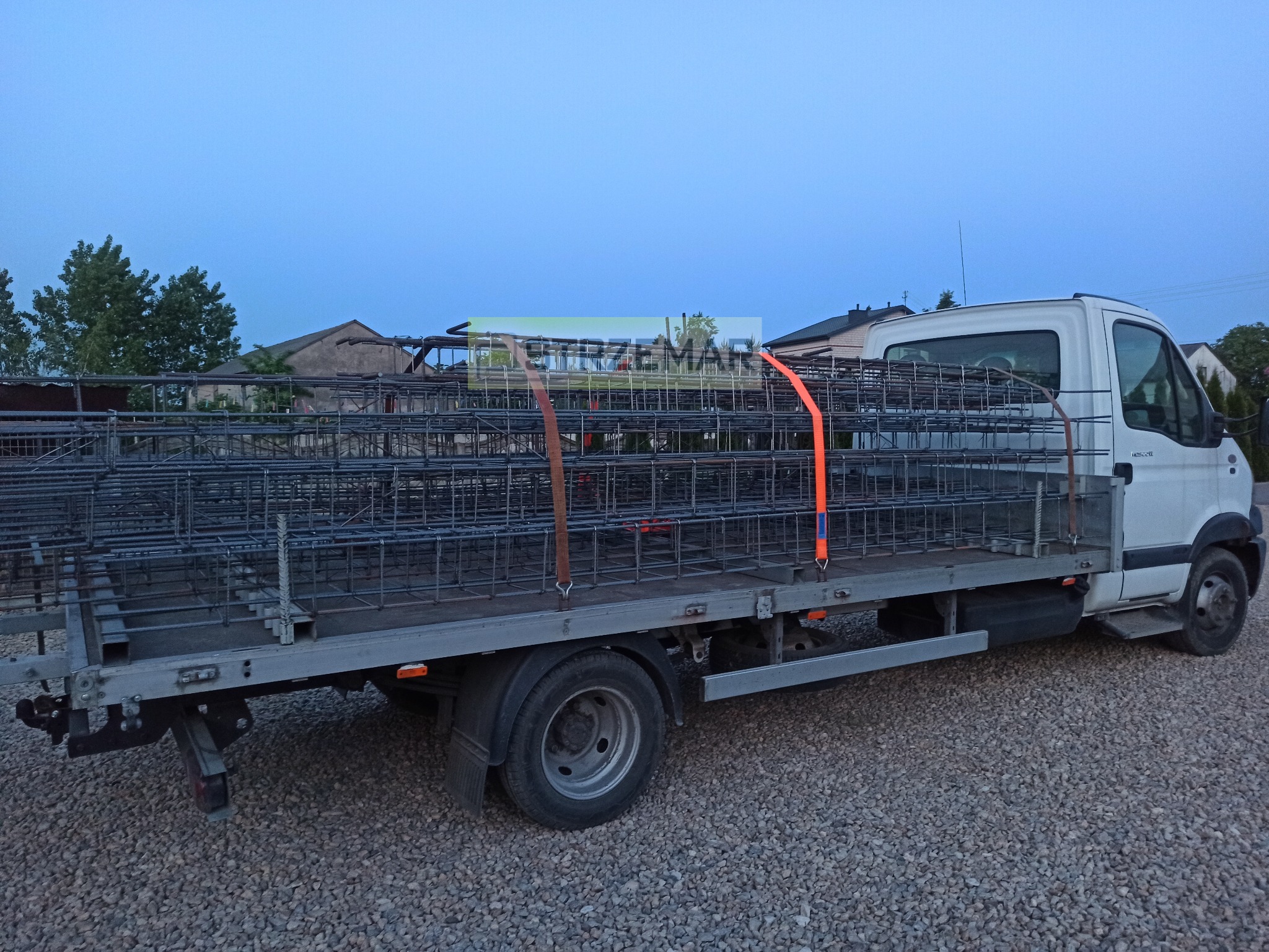 Biały samochód dostawczy z platformą załadowaną siatkami z prętów, zabezpieczonymi pomarańczowymi pasami transportowymi, na żwirowym podłożu.