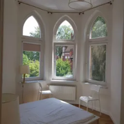 Jasna sypialnia z trzema gotyckimi oknami, białym łóżkiem i krzesłami. Widok na zieleń za oknem. Minimalistyczny wystrój, parkiet na podłodze.