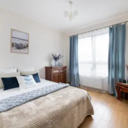 Jasna sypialnia z łóżkiem, beżowo-błękitną narzutą i dekoracjami w stylu marynistycznym. Drewniane meble, niebieskie zasłony, obraz z łodzią.