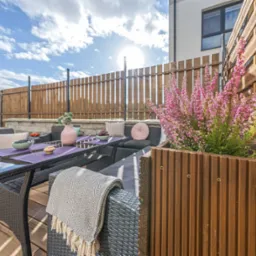 Umeblowany taras z rattanowymi meblami, drewnianą donicą z wrzosem i dekoracjami stołu w fioletach, na tle drewnianego ogrodzenia i błękitnego nieba.