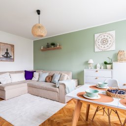 Home Staging Agnieszka Ossowska - Jasne wnętrze z beżową sofą narożną, drewnianym stołem z owocami i zielonymi akcentami. Na ścianie dekoracje w stylu boho i obraz z motywem jogi.