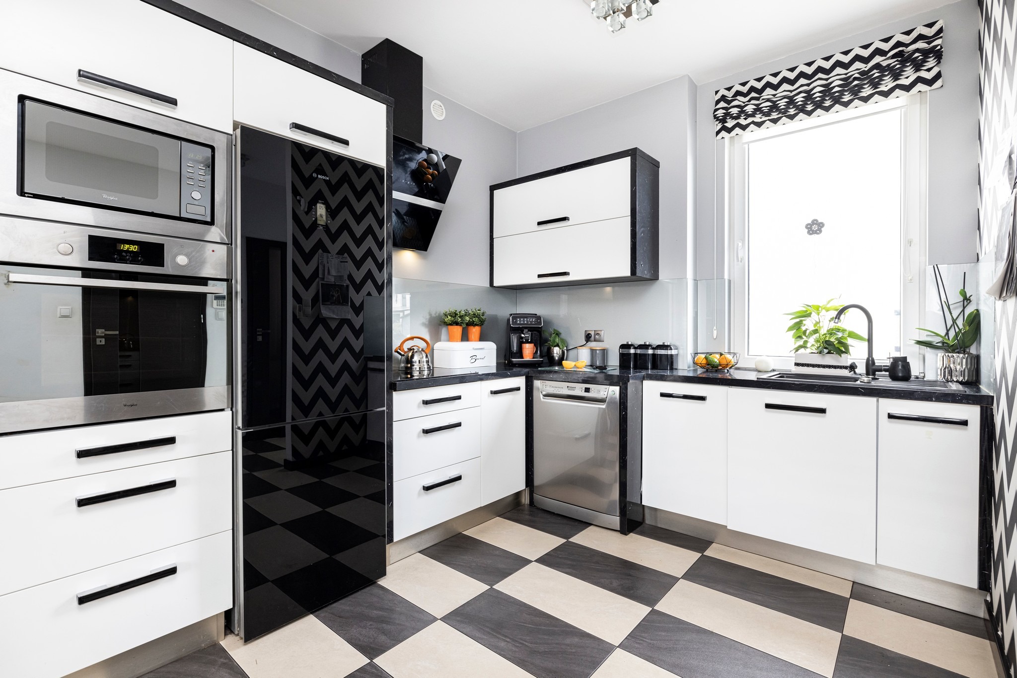 Nowoczesna kuchnia w bieli i czerni: szachownica na podłodze, zabudowa z piekarnikiem i mikrofalówką, okap nad płytą, roleta w zygzaki. Rośliny i owoce jako akcenty.