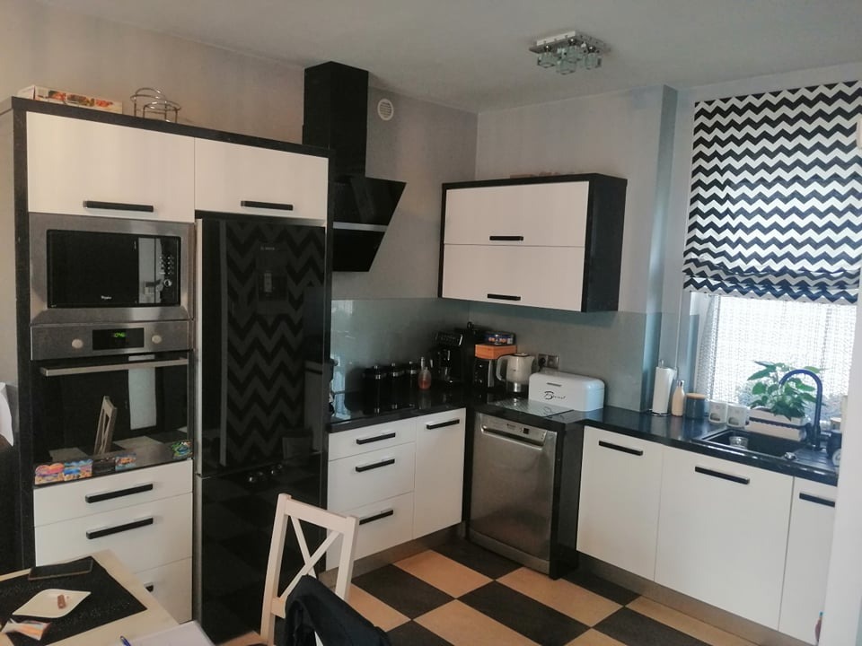 Nowoczesna kuchnia w bieli i czerni: zabudowa z piekarnikiem i mikrofalówką, okap nad płytą, blat z akcesoriami, okno z roletą w zygzaki, szachownica na podłodze.