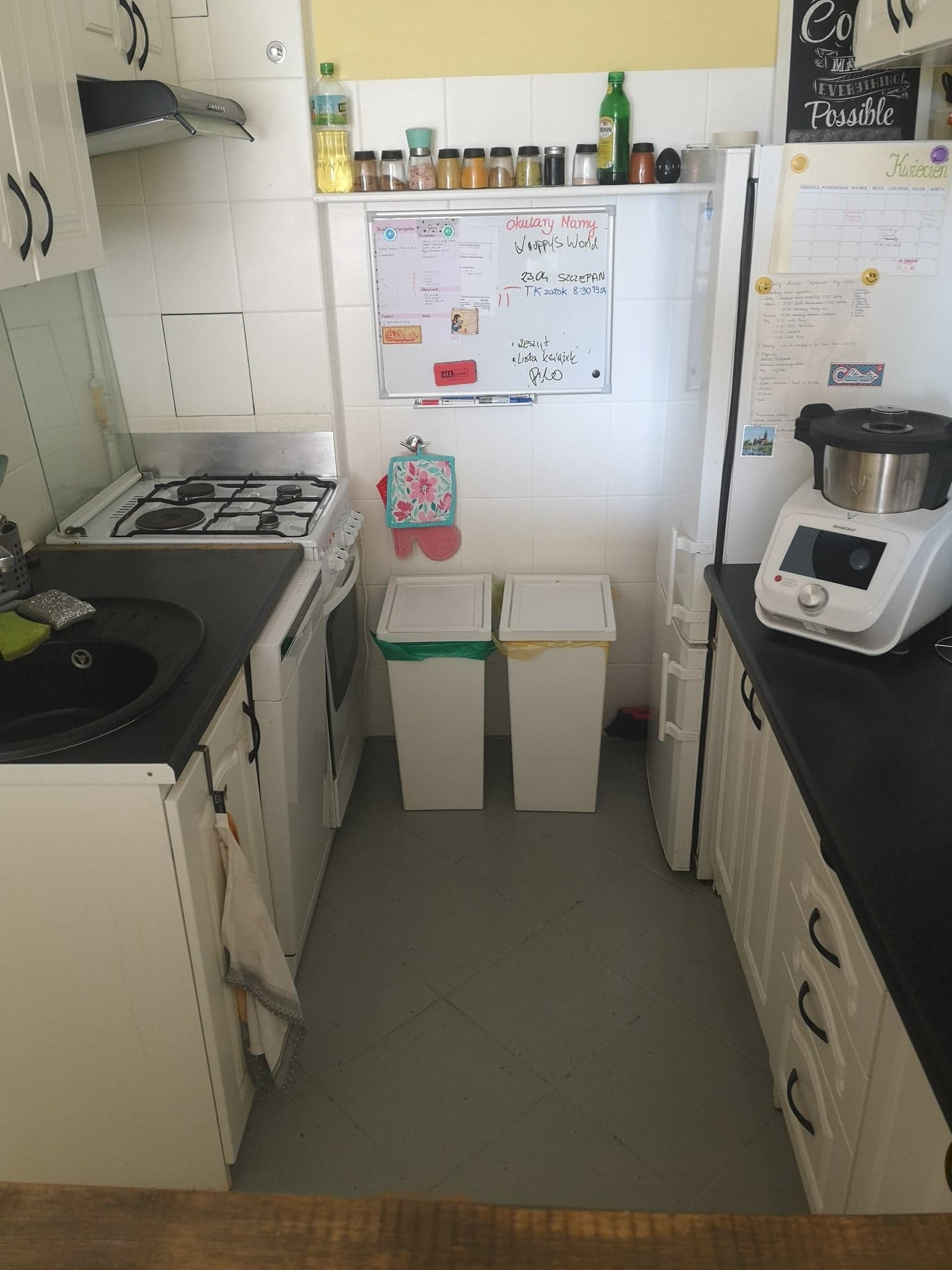Kuchnia z białymi szafkami, czarnym blatem i sprzętem AGD. Na ścianie tablica z notatkami i kalendarz. Dwa kosze na śmieci przy ścianie. Widok z perspektywy osoby stojącej.