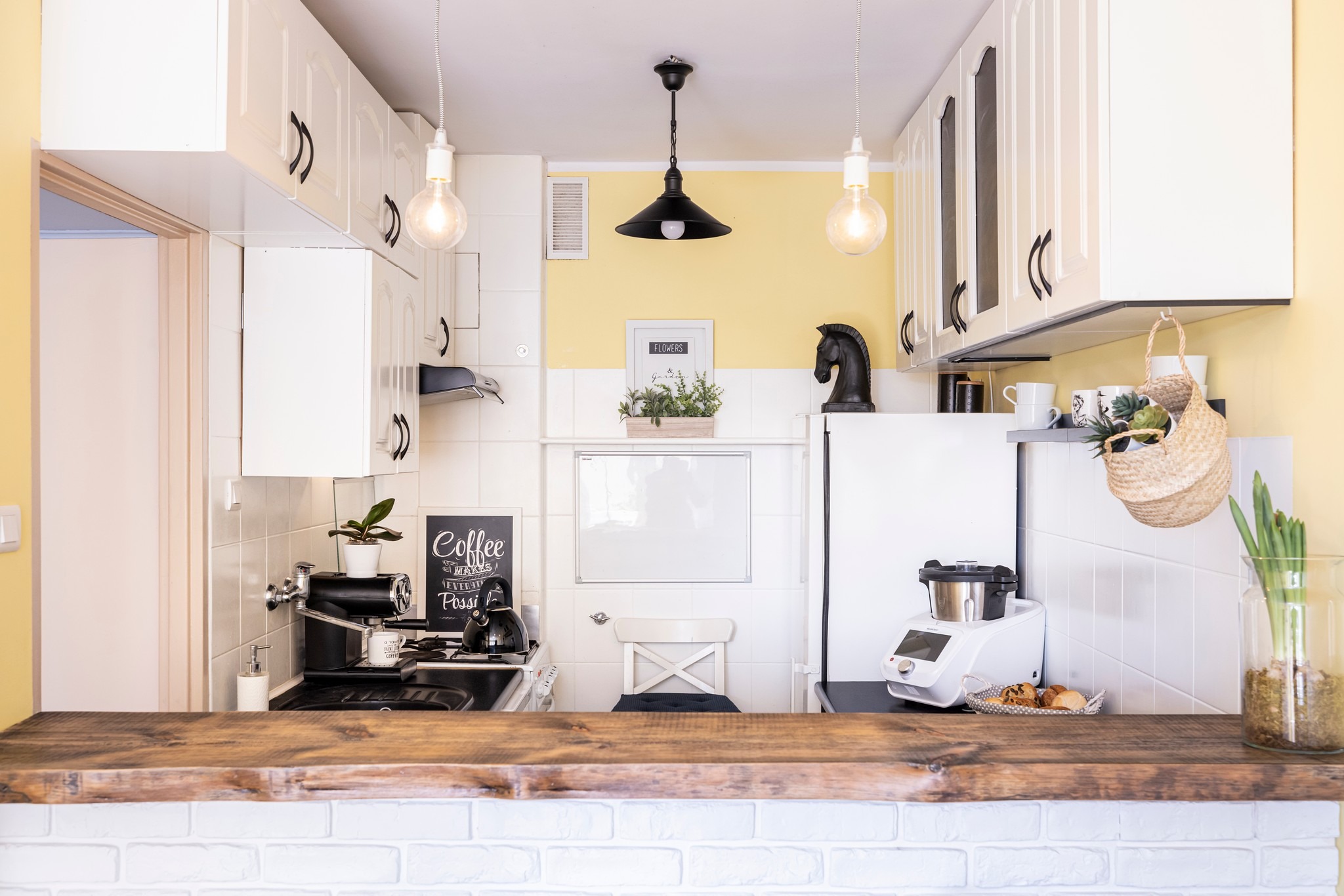 Jasna kuchnia z drewnianym blatem i dekoracjami: tablica kredowa, roślina w doniczce, ozdobny koń. Żółta ściana i białe szafki tworzą przytulną przestrzeń.