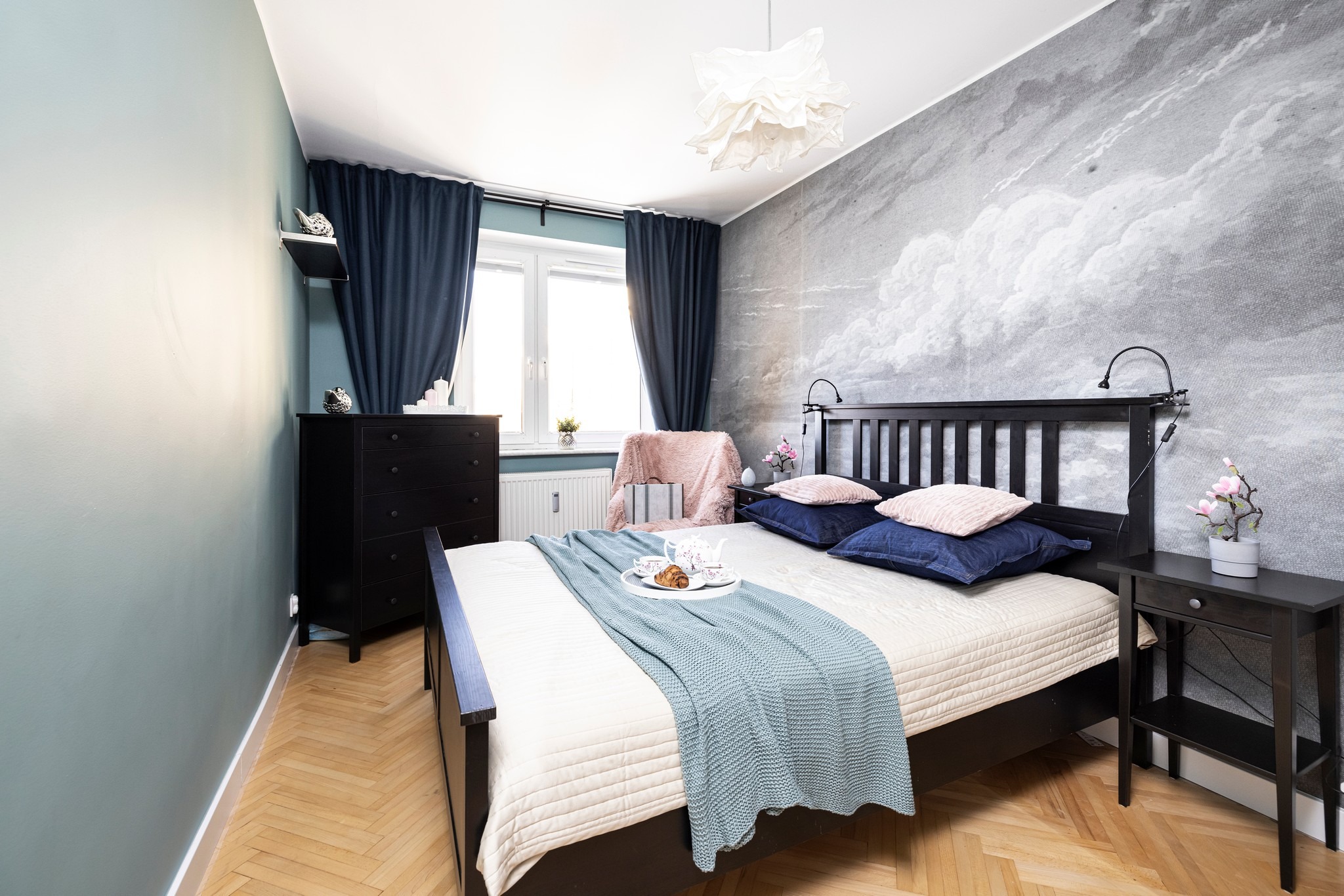 Jasny pokój z łóżkiem, stolikiem nocnym i komodą. Ściana z motywem chmur, niebieskie zasłony i tekstylia. Na łóżku taca z śniadaniem. Przytulna aranżacja.