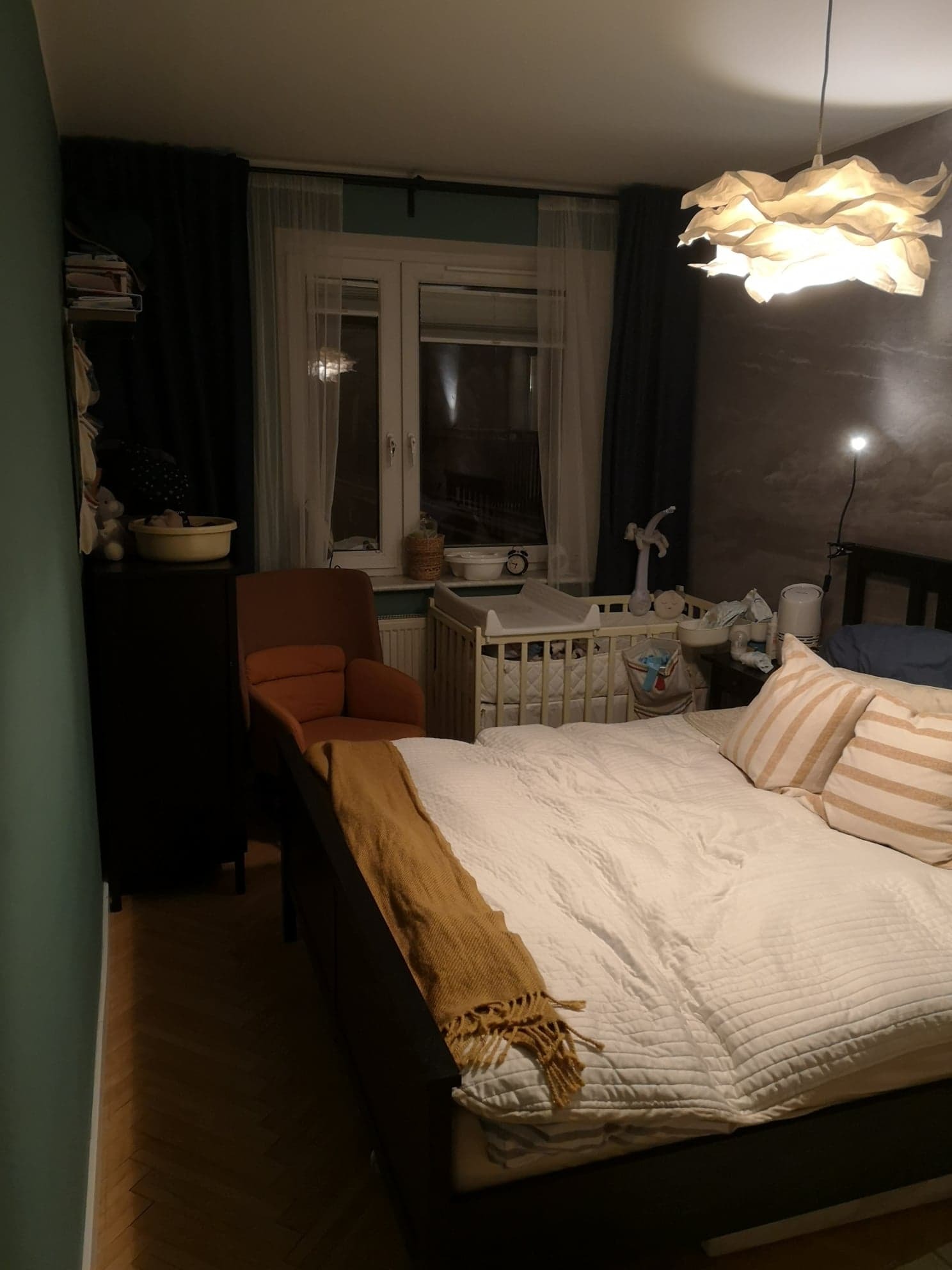 Przytulna sypialnia z łóżkiem, fotelem i komodą w stonowanych barwach. W tle widoczne łóżeczko dziecięce i okno z zasłonami. Ciepłe oświetlenie.