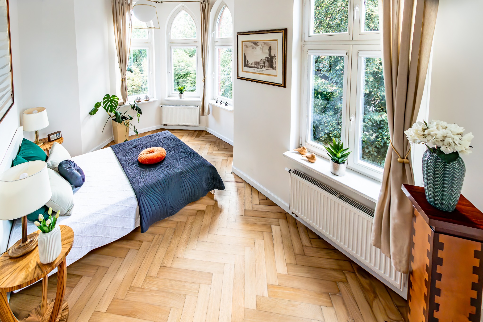 Jasna sypialnia po liftingu: parkiet w jodełkę, łóżko z dekoracyjnymi poduszkami, okna z widokiem na zieleń, stylowe dodatki i kwiaty w wazonie.