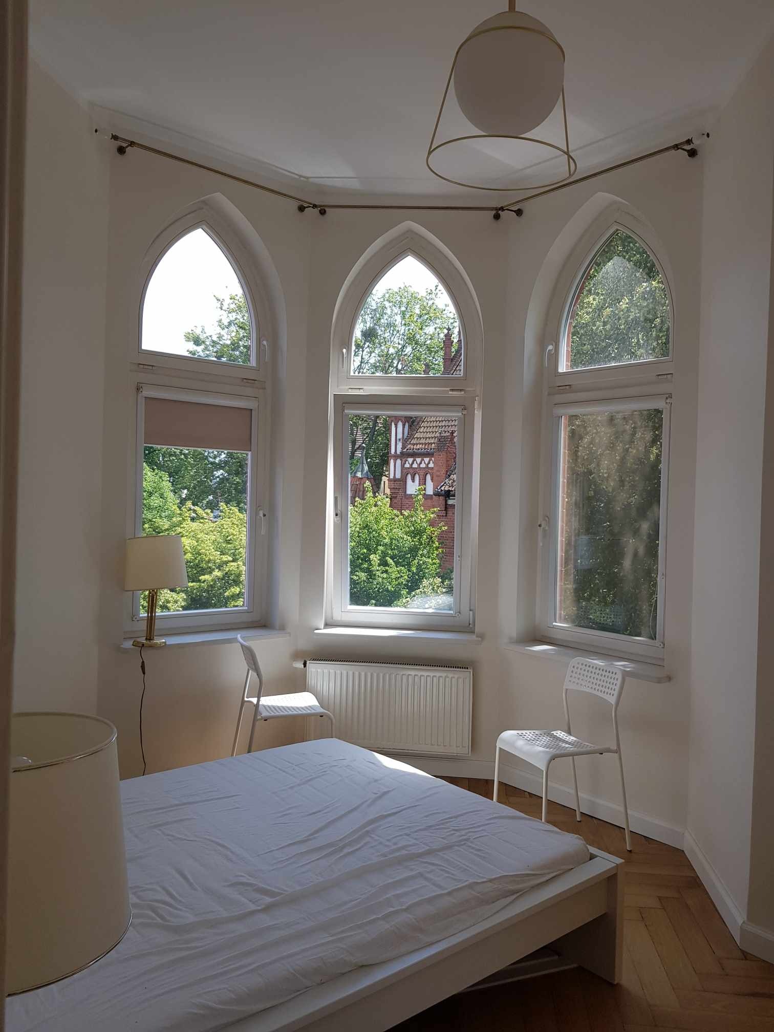 Jasna sypialnia z trzema gotyckimi oknami, białym łóżkiem i krzesłami. Widok na zieleń za oknem. Minimalistyczny wystrój, parkiet na podłodze.