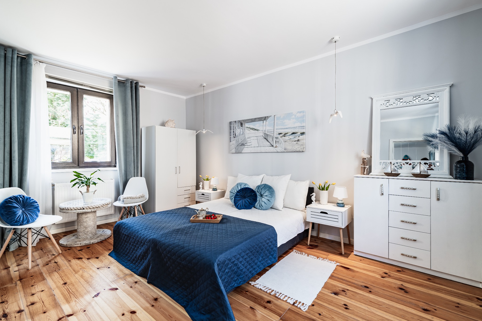 Jasna sypialnia z drewnianą podłogą, niebieską narzutą na łóżku i dekoracjami w stylu marynistycznym. Widoczne białe meble i okno z zasłonami.