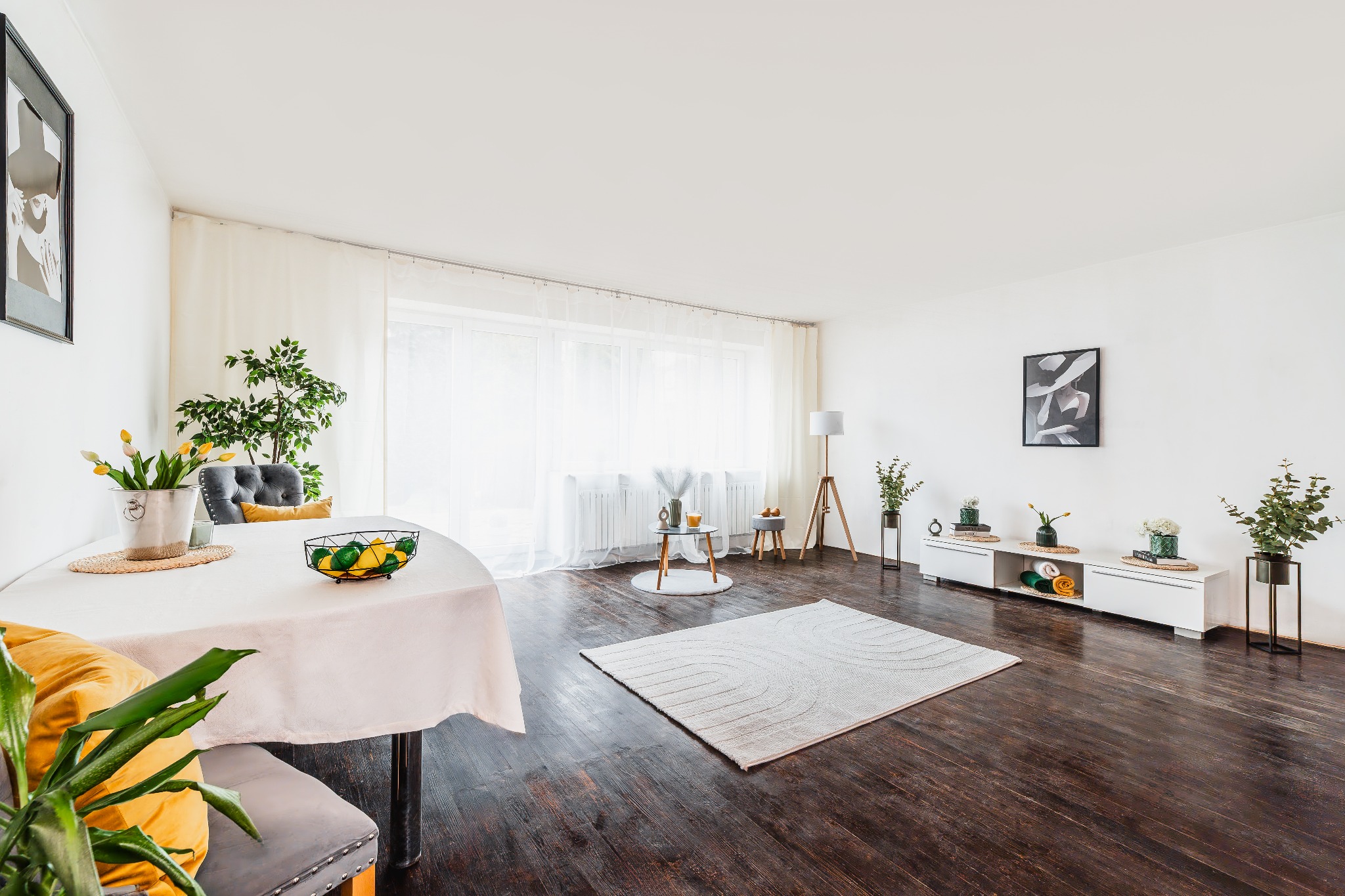 Jasne wnętrze salonu z drewnianą podłogą. Stół z owocami, dywan i biała komoda tworzą przytulną przestrzeń. Minimalistyczny wystrój z akcentami roślinnymi.