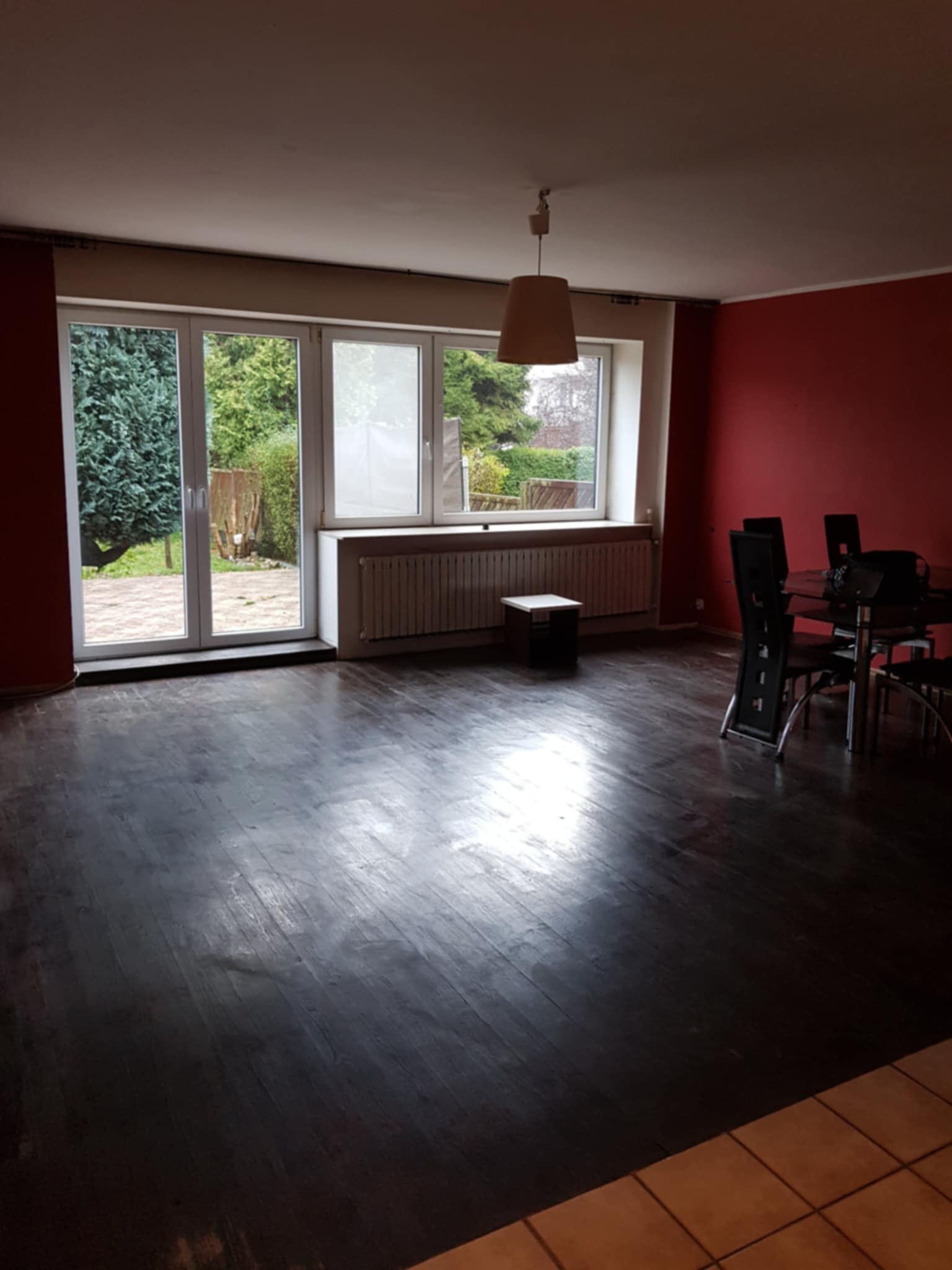 Pusty pokój z ciemną podłogą i czerwonymi ścianami, widok na ogród przez okno balkonowe. Stół z krzesłami i mała szafka przy grzejniku.