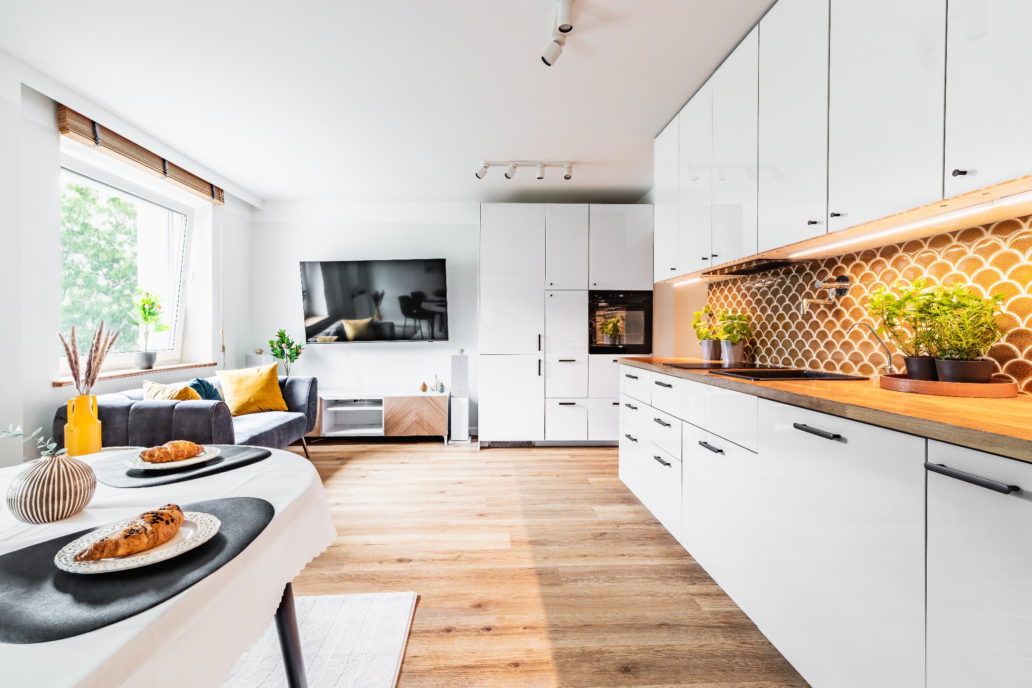 Jasne, przestronne wnętrze z białymi meblami kuchennymi i drewnianym blatem, salon z szarą sofą i stolikiem z croissantami, doświetlone naturalnym światłem.