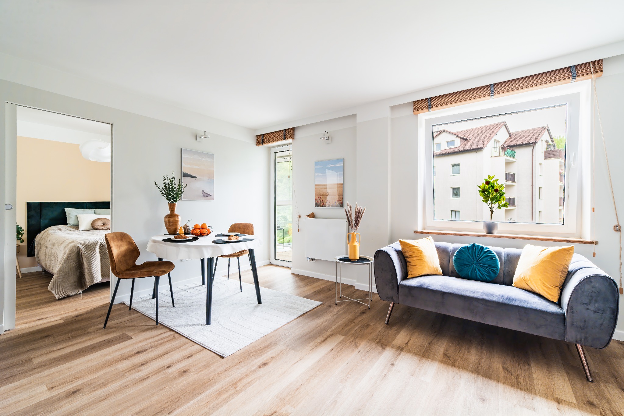 Jasne wnętrze: stół z owocami, szary welurowy narożnik z poduszkami, widok na sypialnię. Roślina w doniczce na parapecie, drewniane żaluzje.