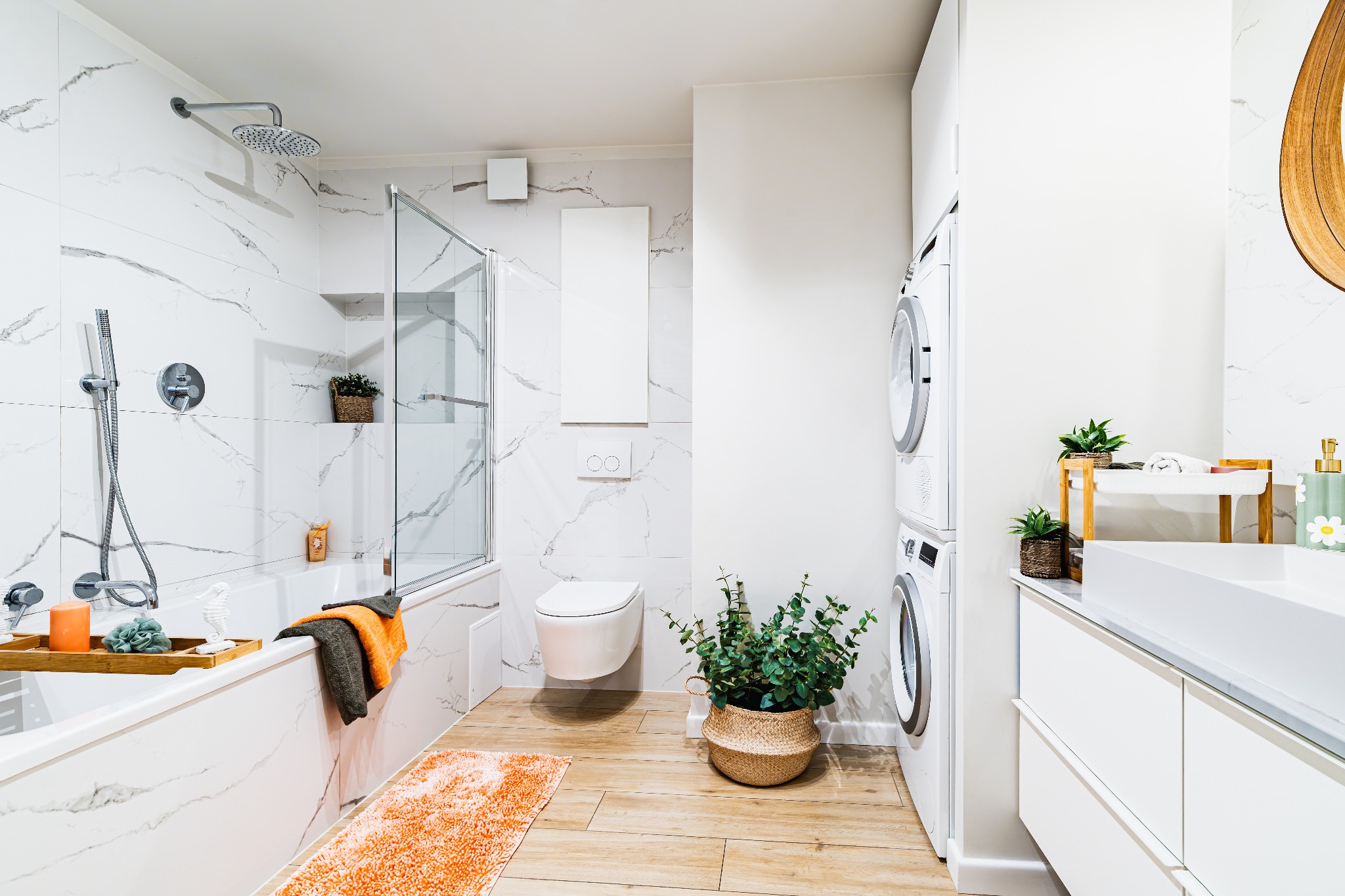 Jasna łazienka z marmurowymi płytkami, drewnianą podłogą i roślinami doniczkowymi. Wanna z parawanem, pralka i suszarka w zabudowie, minimalistyczna umywalka.