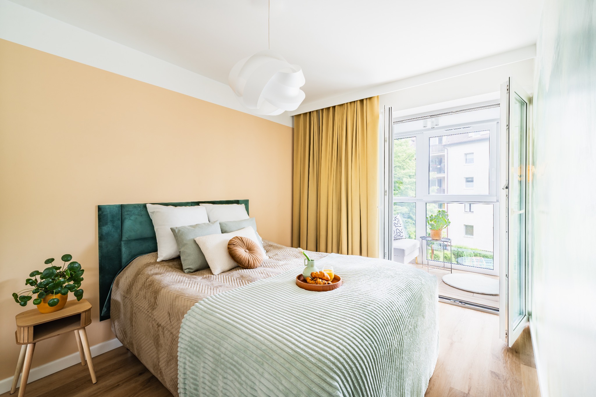 Jasna sypialnia z balkonem: zielony zagłówek, wzorzysta narzuta, śniadanie na łóżku i roślina na stoliku nocnym. Naturalne światło, minimalistyczny wystrój.