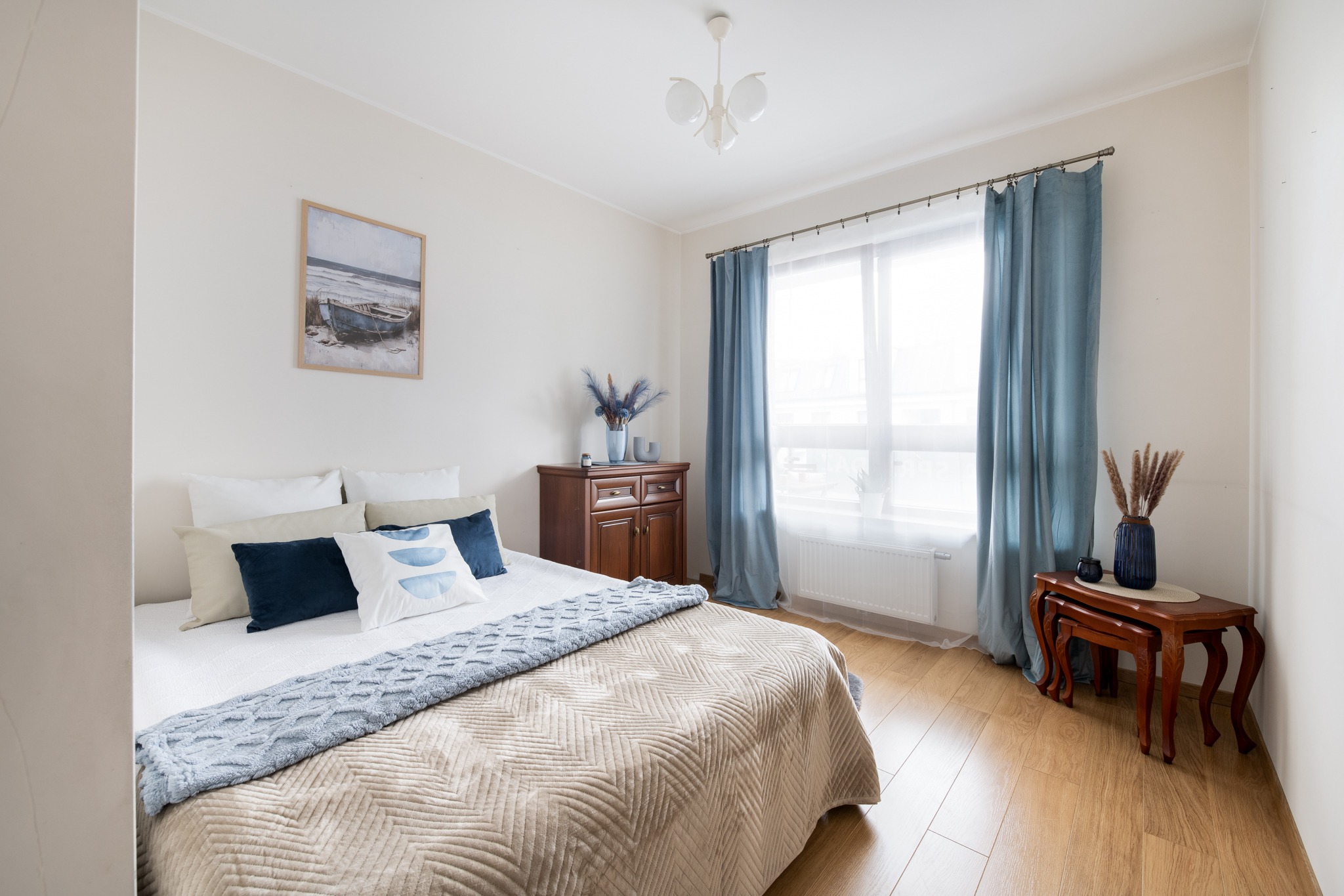 Jasna sypialnia z łóżkiem, beżowo-błękitną narzutą i dekoracjami w stylu marynistycznym. Drewniane meble, niebieskie zasłony, obraz z łodzią.