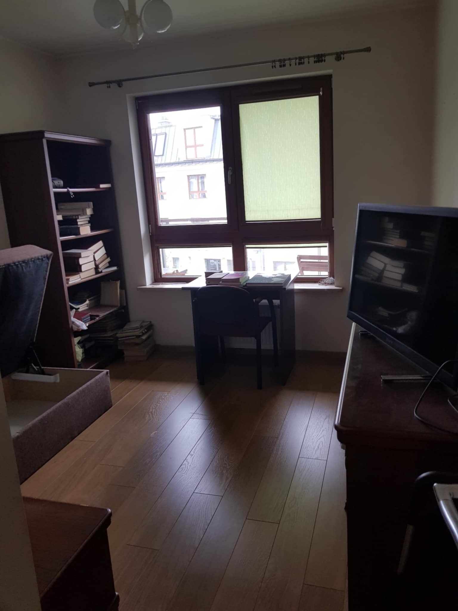 Pokój z drewnianą podłogą, regałem z książkami i biurkiem przy oknie z roletą. Widoczny telewizor na komodzie. Ustawienie mebli przed aranżacją.