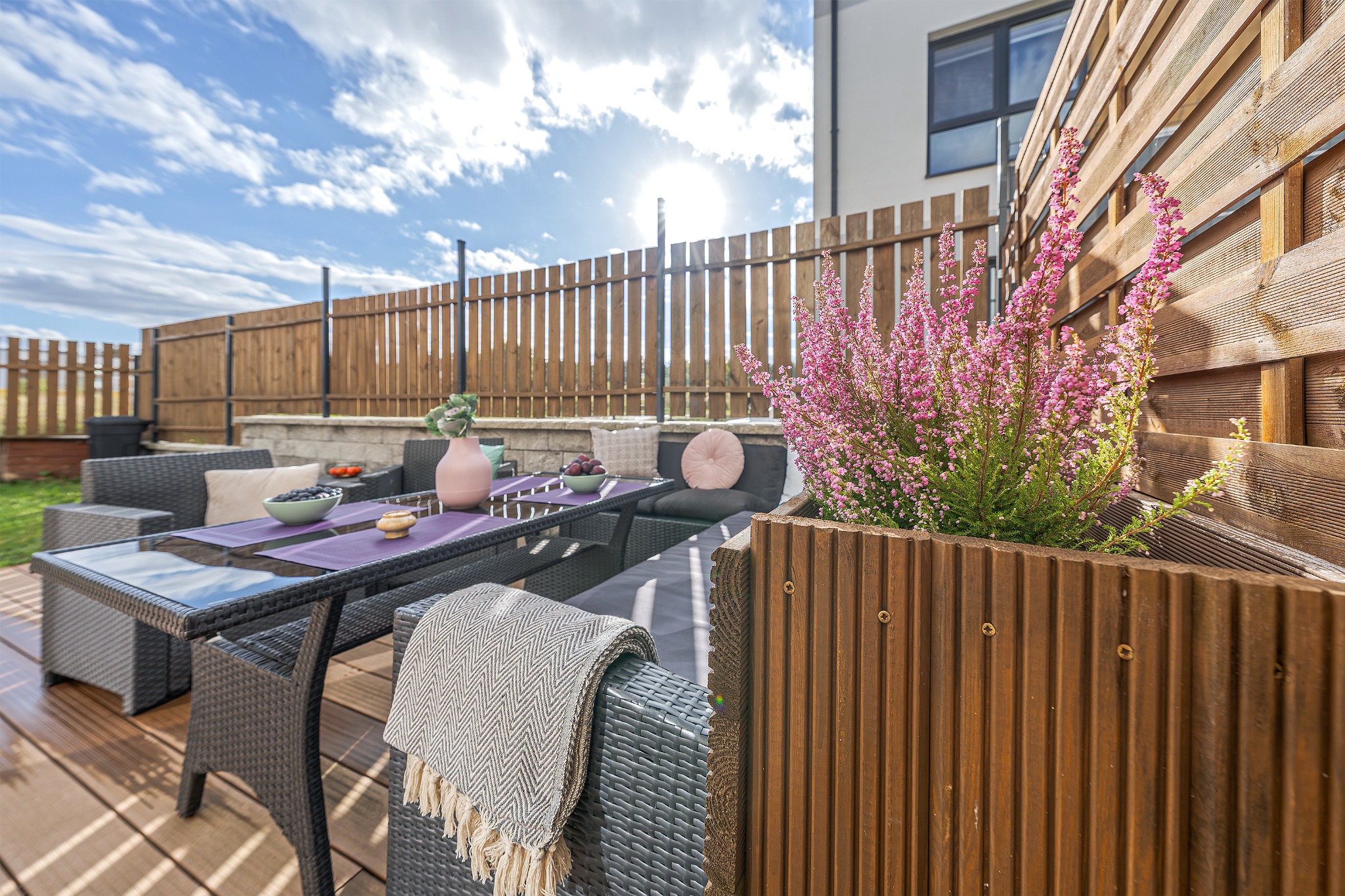 Umeblowany taras z rattanowymi meblami, drewnianą donicą z wrzosem i dekoracjami stołu w fioletach, na tle drewnianego ogrodzenia i błękitnego nieba.