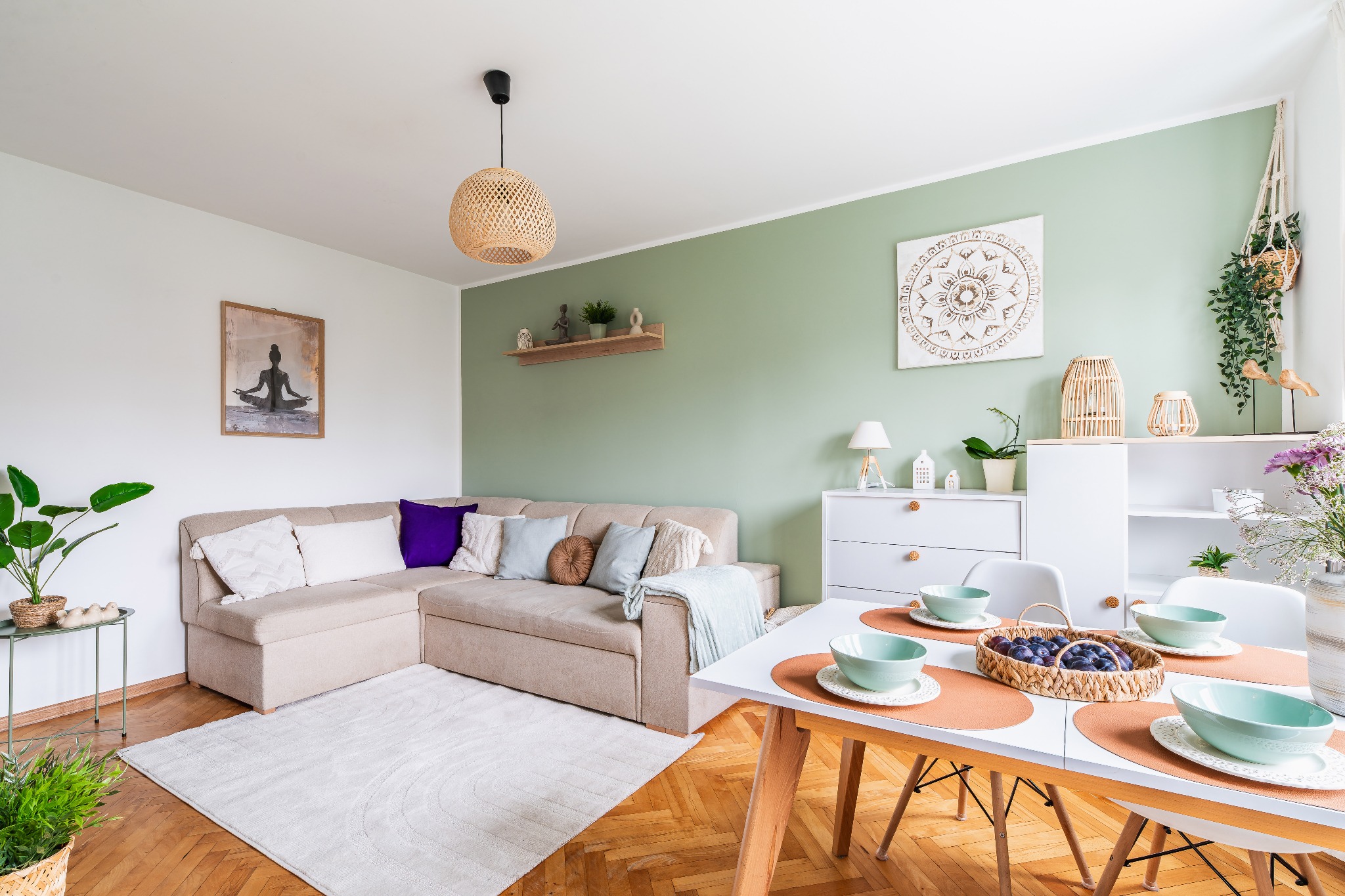 Jasne wnętrze z beżową sofą narożną, drewnianym stołem z owocami i zielonymi akcentami. Na ścianie dekoracje w stylu boho i obraz z motywem jogi.
