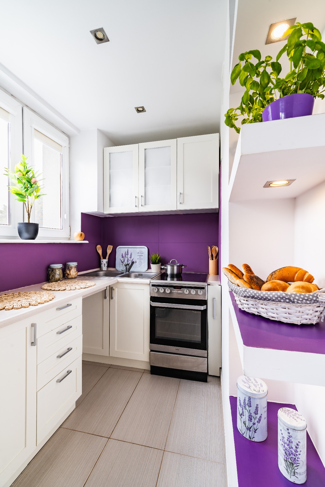 Jasna kuchnia z fioletowymi akcentami: ściana, doniczki, półki. Koszyk z pieczywem i zioła dodają przytulności. Nowoczesny design z dbałością o detale.