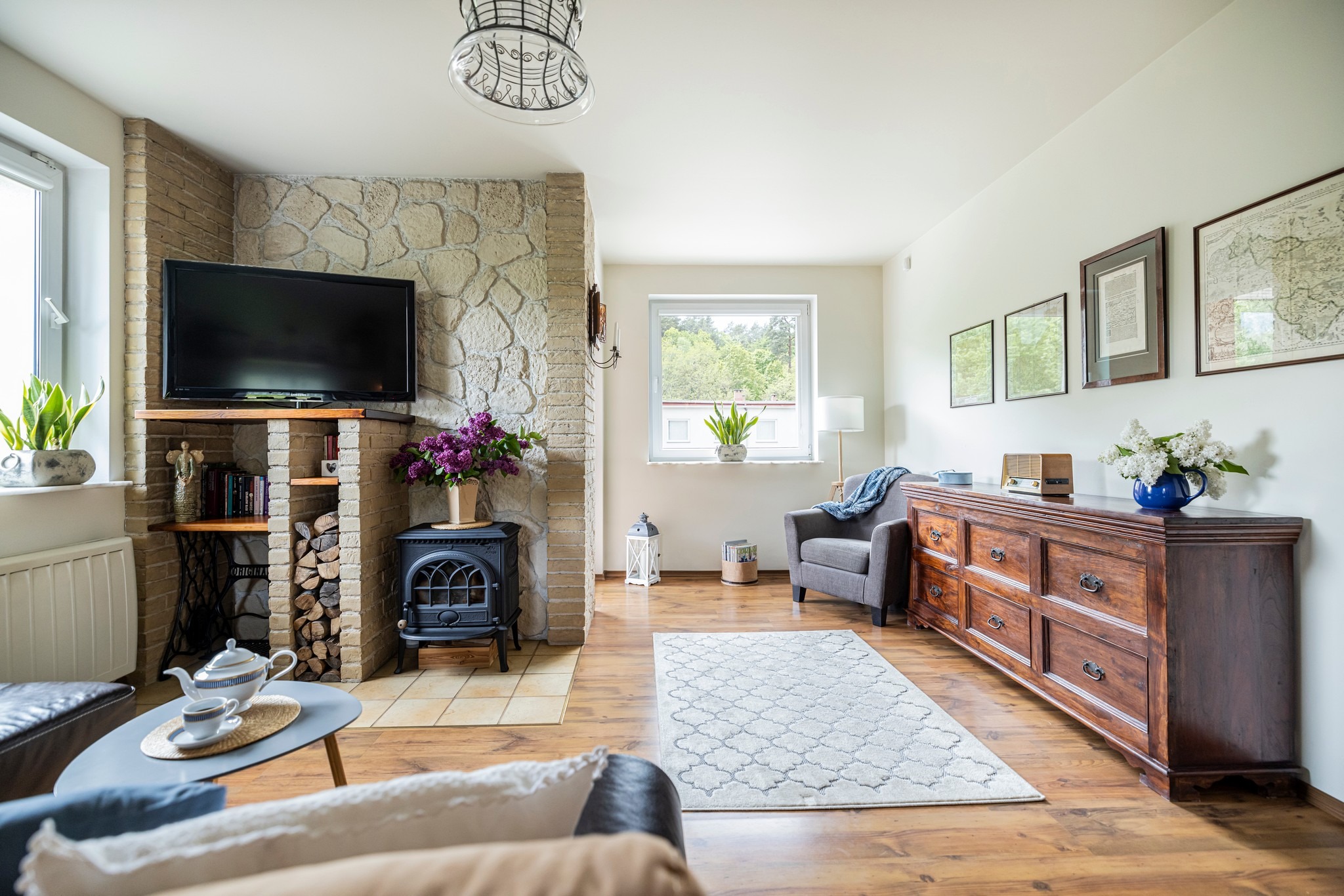 Jasne wnętrze salonu z kamiennym kominkiem, drewnianą komodą i szarym fotelem. Na pierwszym planie stolik z serwisem do herbaty. Przytulna aranżacja.