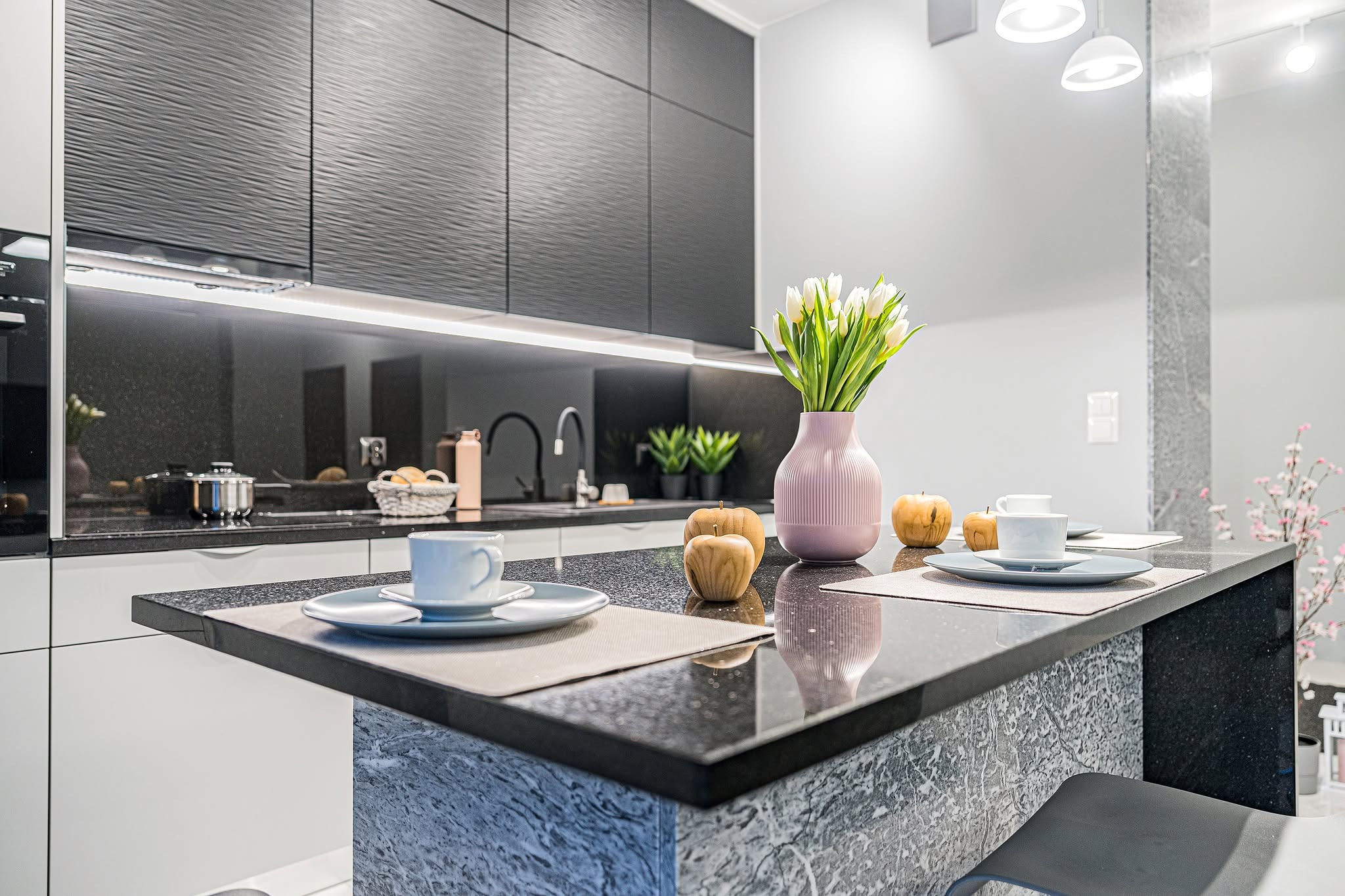 Nowoczesna kuchnia z czarnym blatem i frontami szafek o falistej strukturze. Na blacie wazon z tulipanami, jabłka i filiżanki. Stonowana kolorystyka, minimalistyczny design.