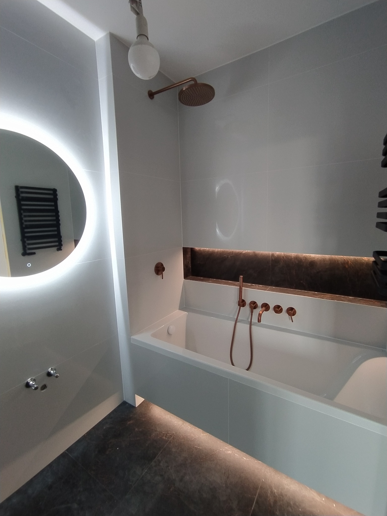 Nowoczesna łazienka z wanną, armaturą w kolorze miedzi i okrągłym lustrem z podświetleniem LED.