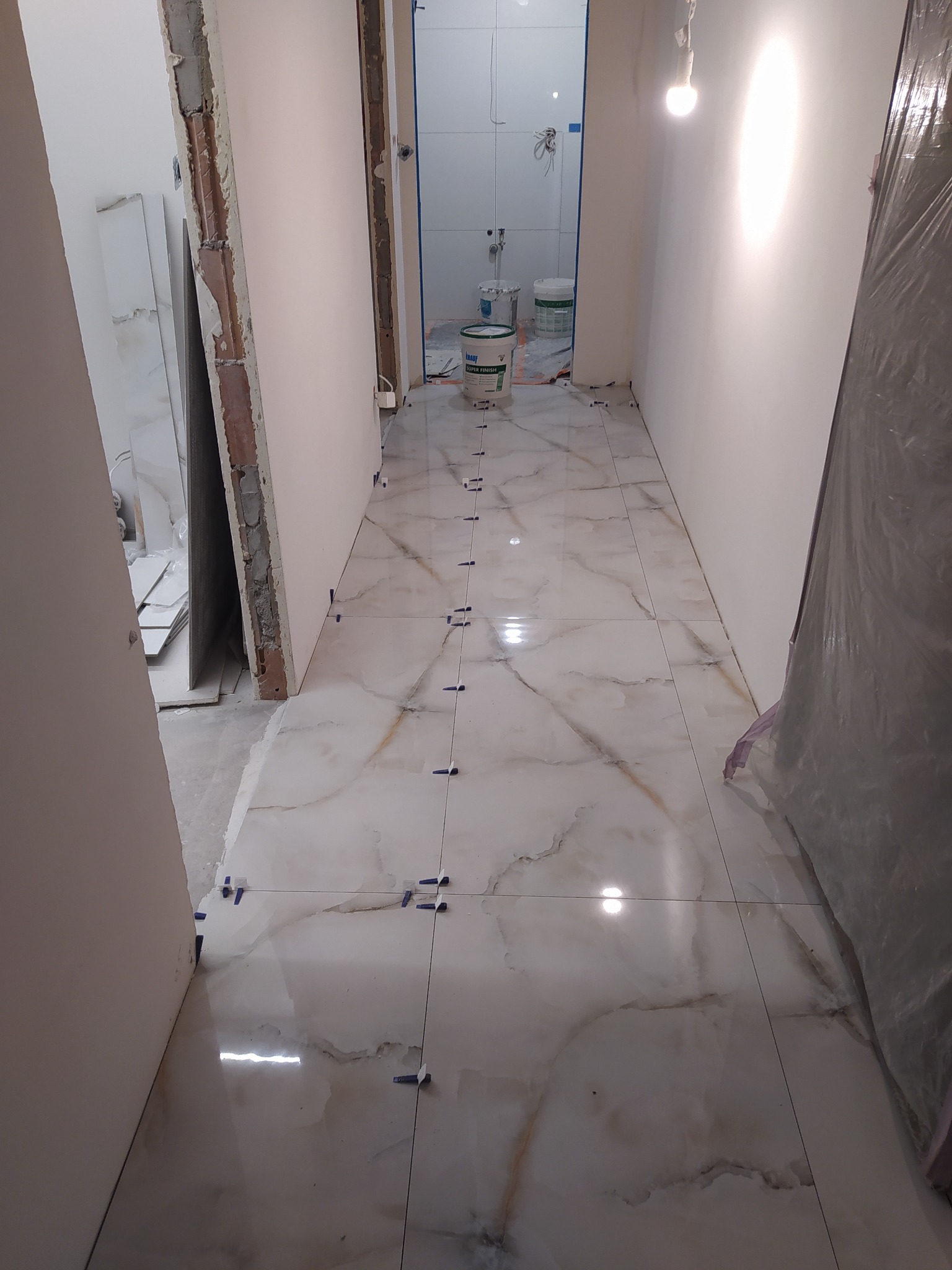 Układanie błyszczących płytek podłogowych z marmurowym wzorem w trakcie prac wykończeniowych łazienki, widoczne krzyżyki dystansowe i puste ściany przygotowane do malowania.