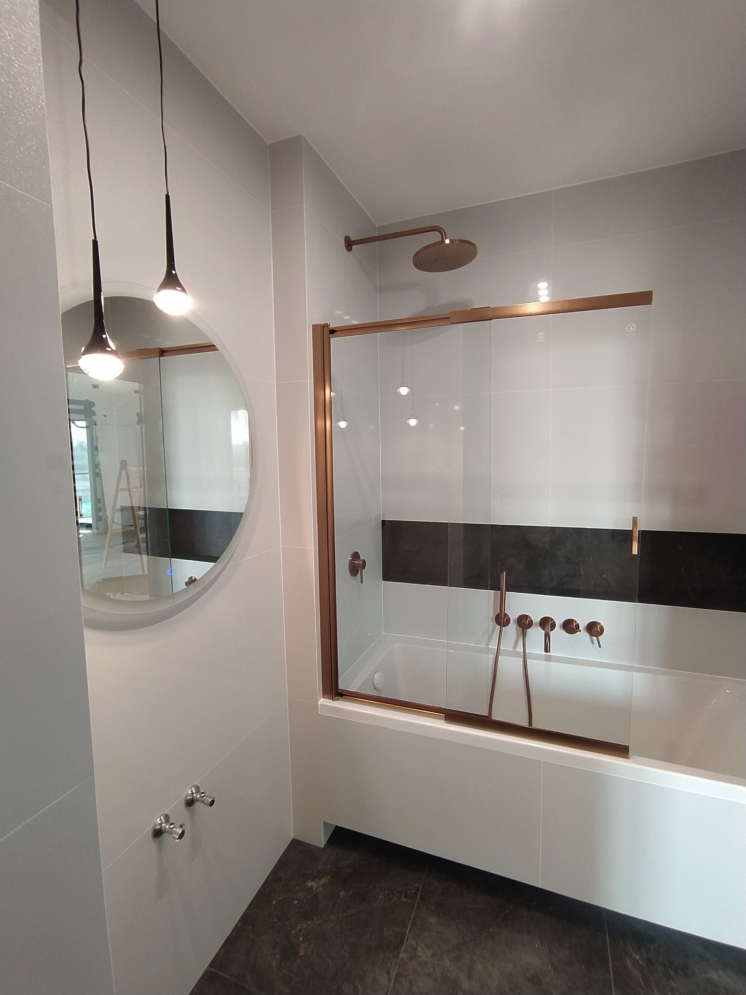Nowoczesna łazienka z wanną za szklaną ścianką z miedzianymi ramami, miedziana deszczownica, armatura w kolorze miedzi, okrągłe lustro i wiszące lampy.