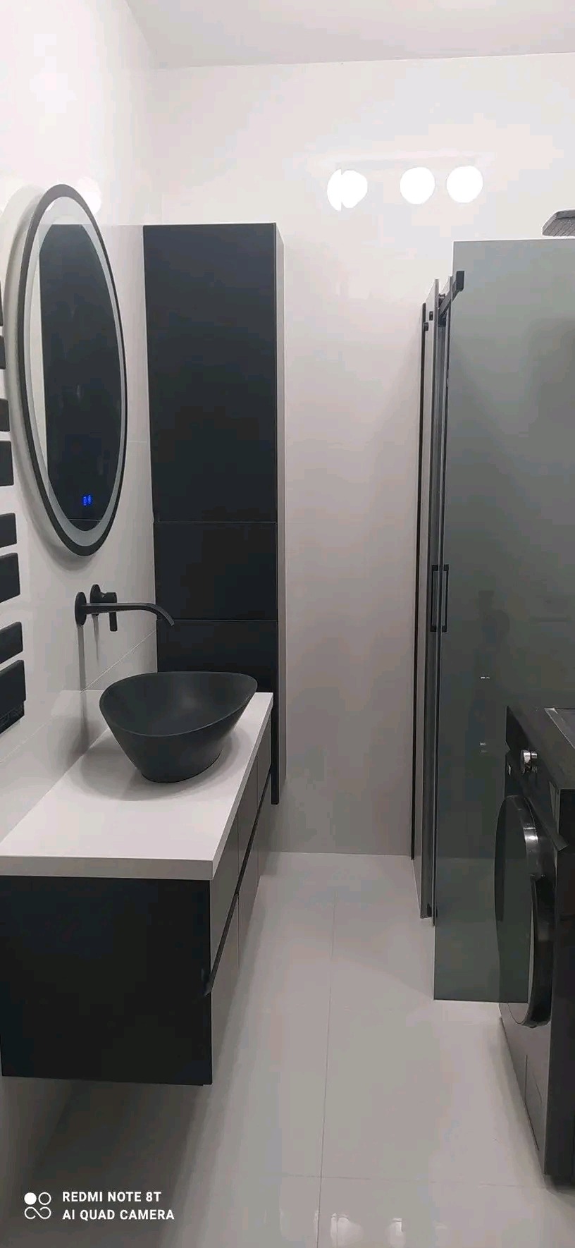 Nowoczesna łazienka z czarną, owalną umywalką na białym blacie, czarną armaturą i podświetlanym lustrem, obok kabina prysznicowa i pralka.