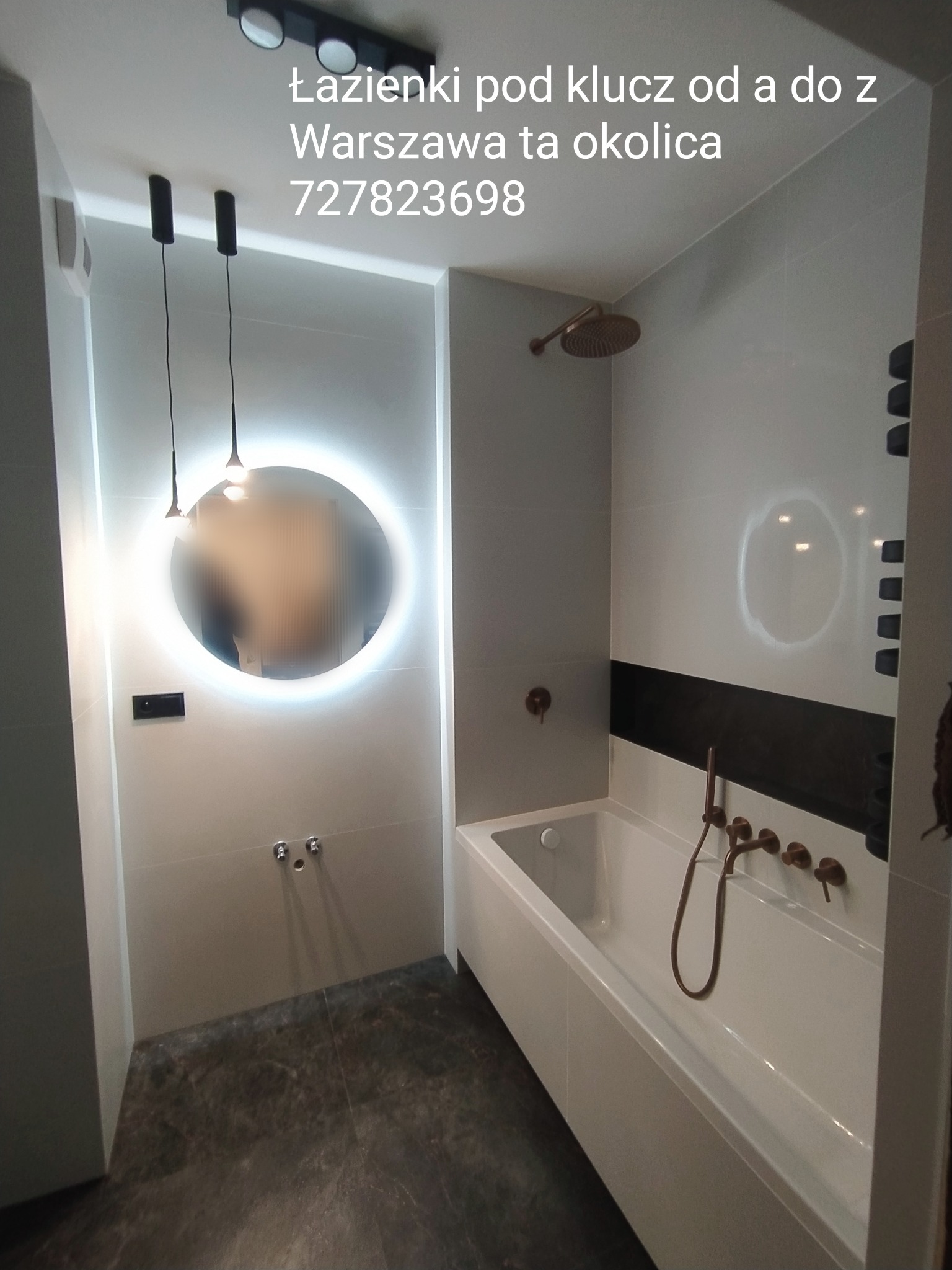 Nowoczesna łazienka z podświetlanym okrągłym lustrem, deszczownicą, wanną i ciemną podłogą. Armatura w kolorze miedzianym.