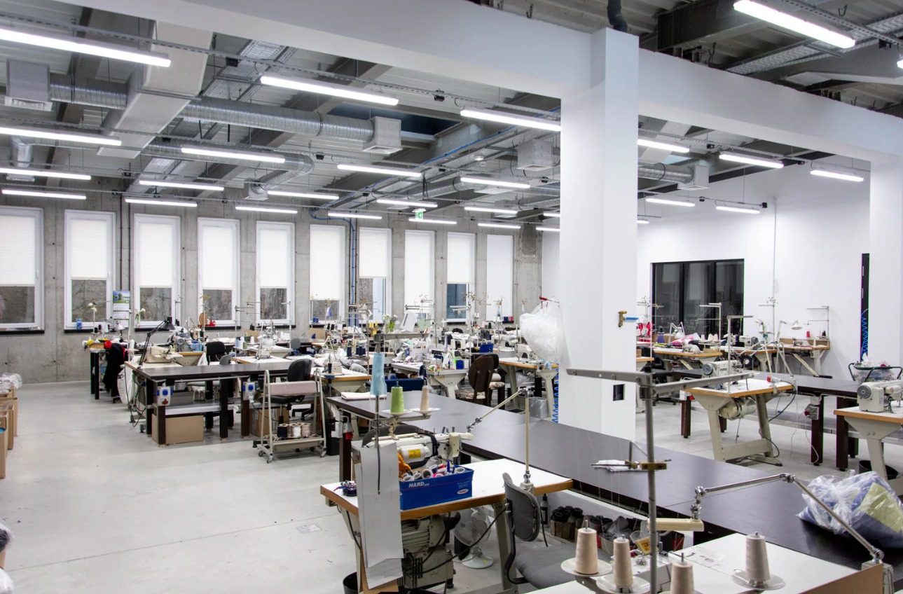 Przestronna, jasna szwalnia z rzędami stanowisk roboczych wyposażonych w maszyny do szycia, liczne okna z roletami i przemysłowe oświetlenie sufitowe.