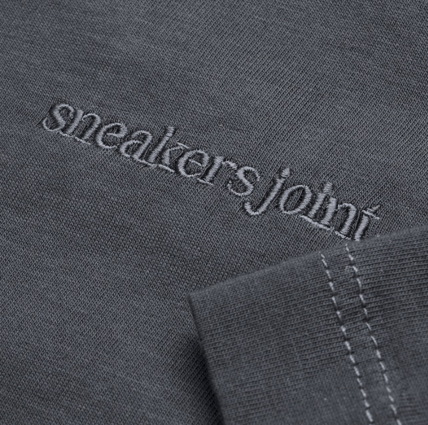 Szara koszulka z wyhaftowanym napisem 'sneakersjoint' w dolnym rogu, widoczne szwy na rękawie.