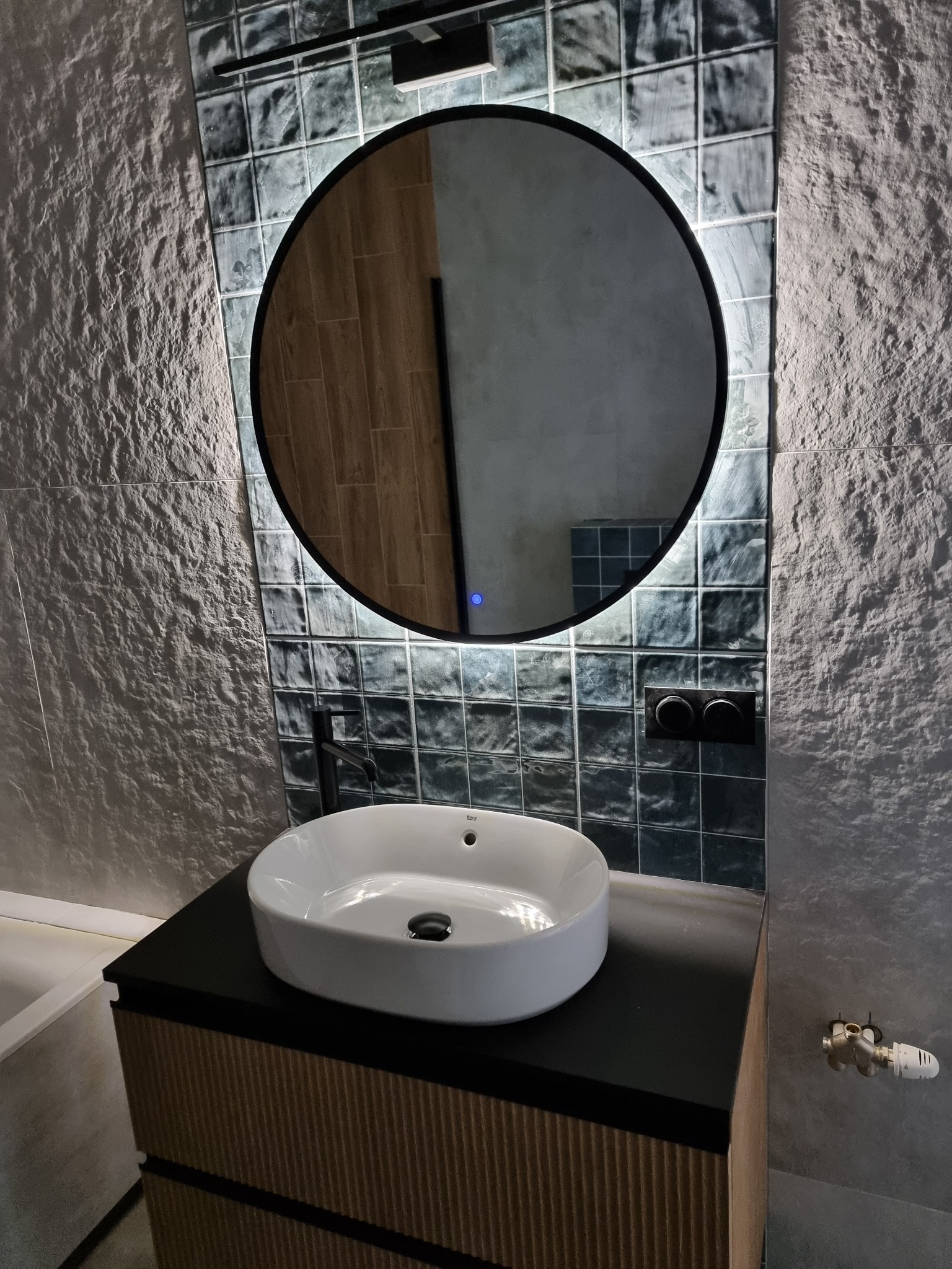 Nowoczesna łazienka z umywalką nablatową, okrągłym lustrem z podświetleniem LED i dekoracyjnymi płytkami imitującymi kamień na ścianie.