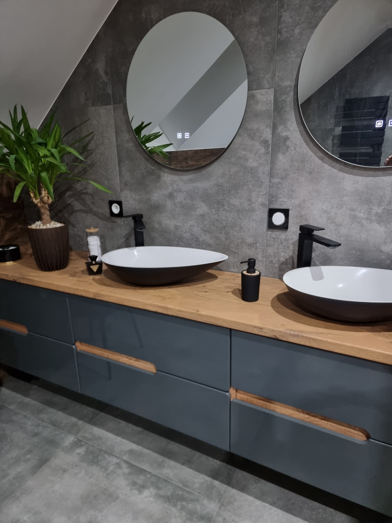 Nowoczesna łazienka z dwoma okrągłymi lustrami z podświetleniem LED, drewnianym blatem i szarą szafką. Uwagę zwracają czarne baterie umywalkowe i minimalistyczny styl.