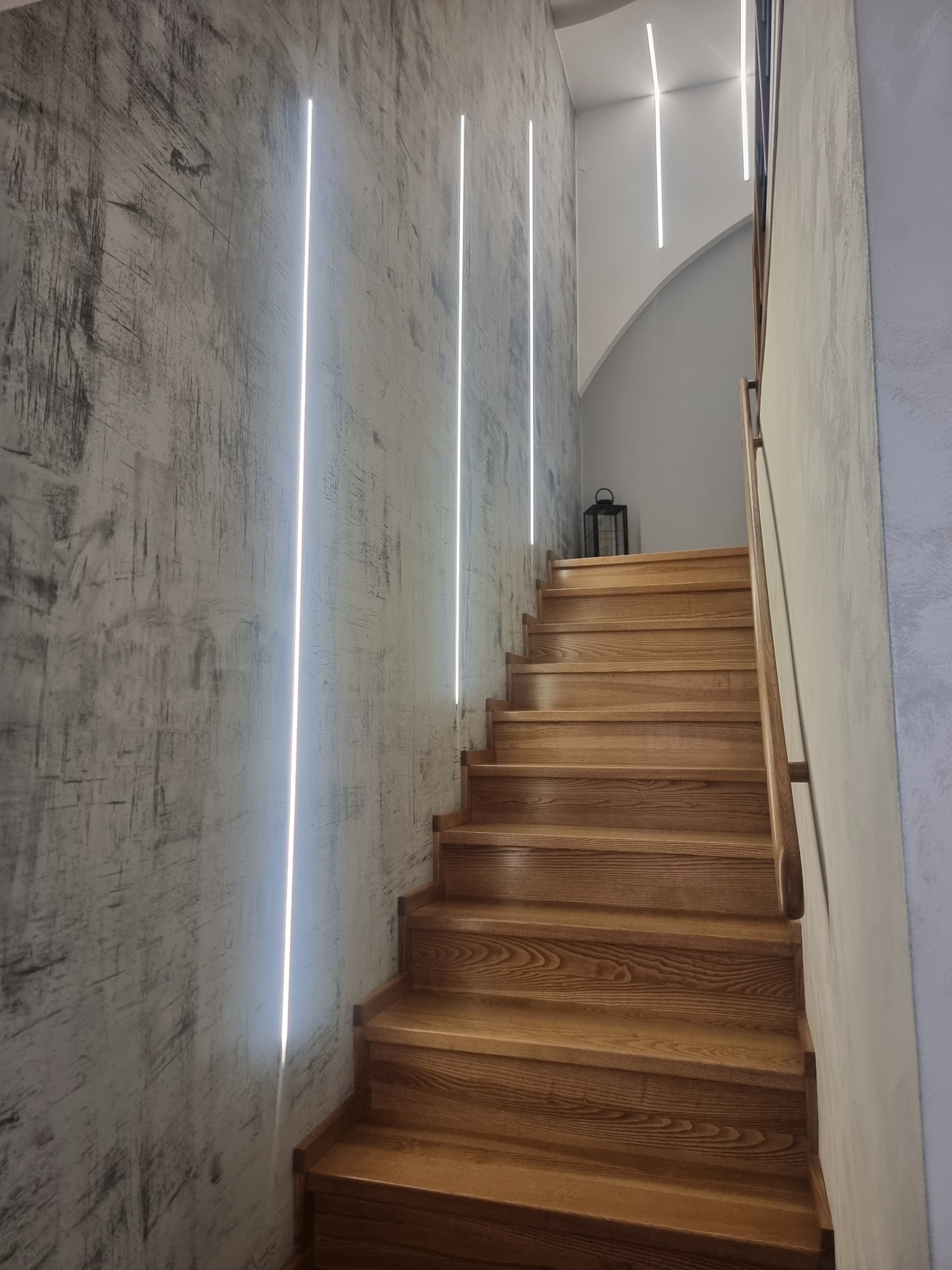 Nowoczesne schody drewniane z oświetleniem LED na ścianie o fakturze betonu. Minimalistyczny design, ciepłe drewno kontrastuje z chłodnym odcieniem ściany.