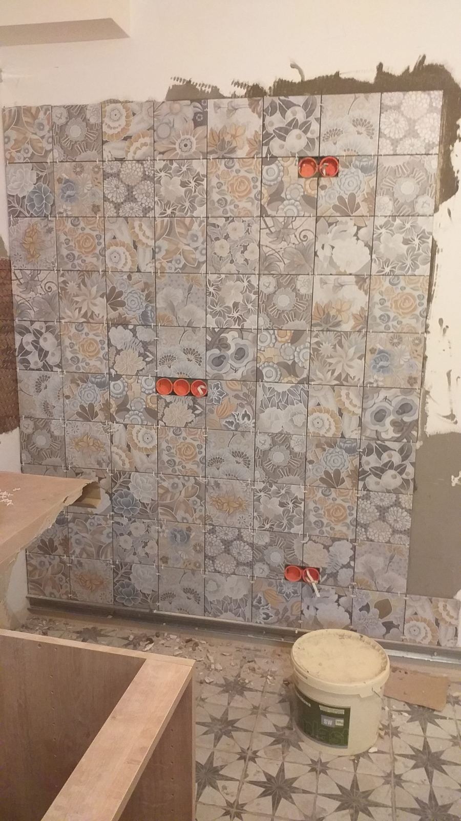 Układanie dekoracyjnych płytek ściennych z motywem kwiatowym w łazience w Warszawie. Widoczne puszki elektryczne i wiadro z zaprawą.