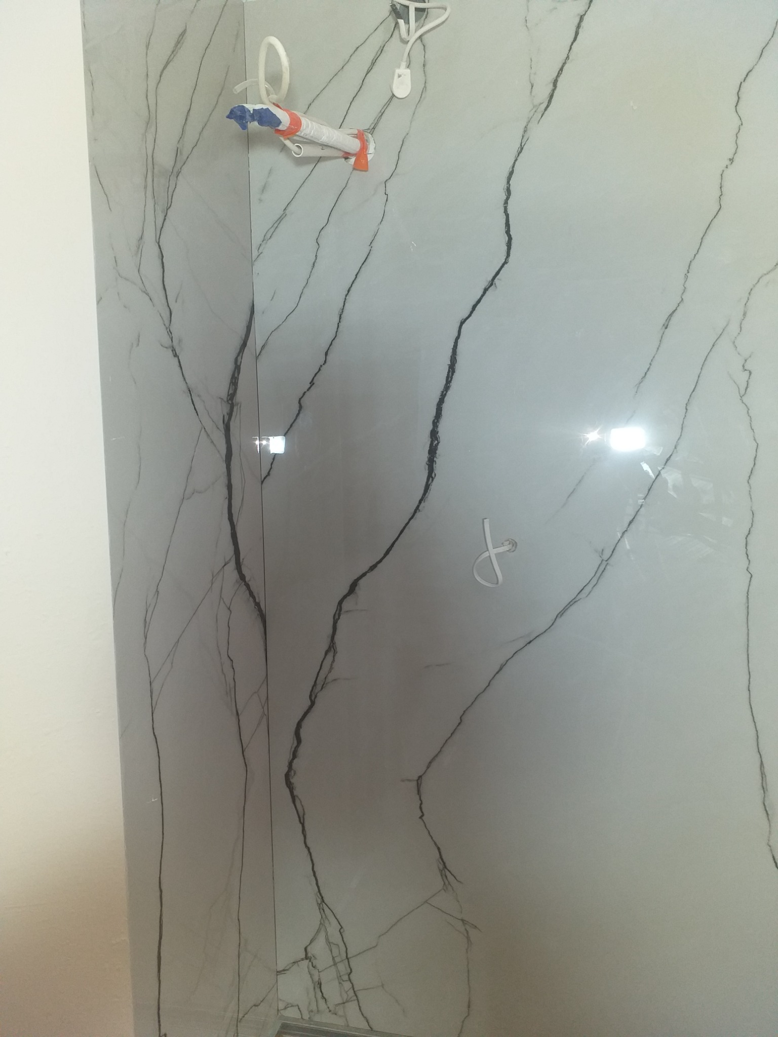 Ściana wyłożona błyszczącymi, szarymi płytkami imitującymi marmur z wyraźnym, ciemnym wzorem żyłkowania. Widoczne akcesoria montażowe i fragment sąsiedniej ściany.