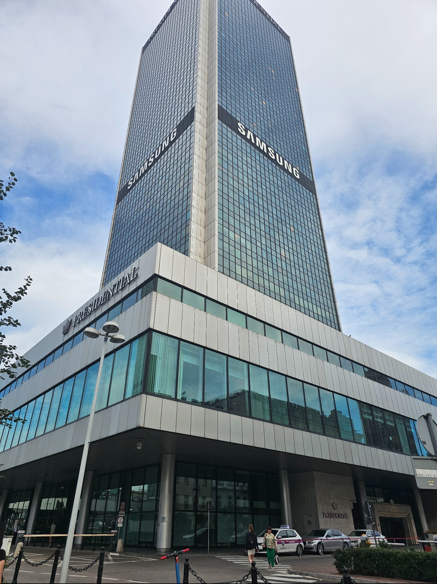 Wysoki biurowiec z napisem SAMSUNG na tle błękitnego nieba. Budynek Presidential z nowoczesną architekturą i szklaną fasadą. Widok z dołu, podkreślający monumentalność.