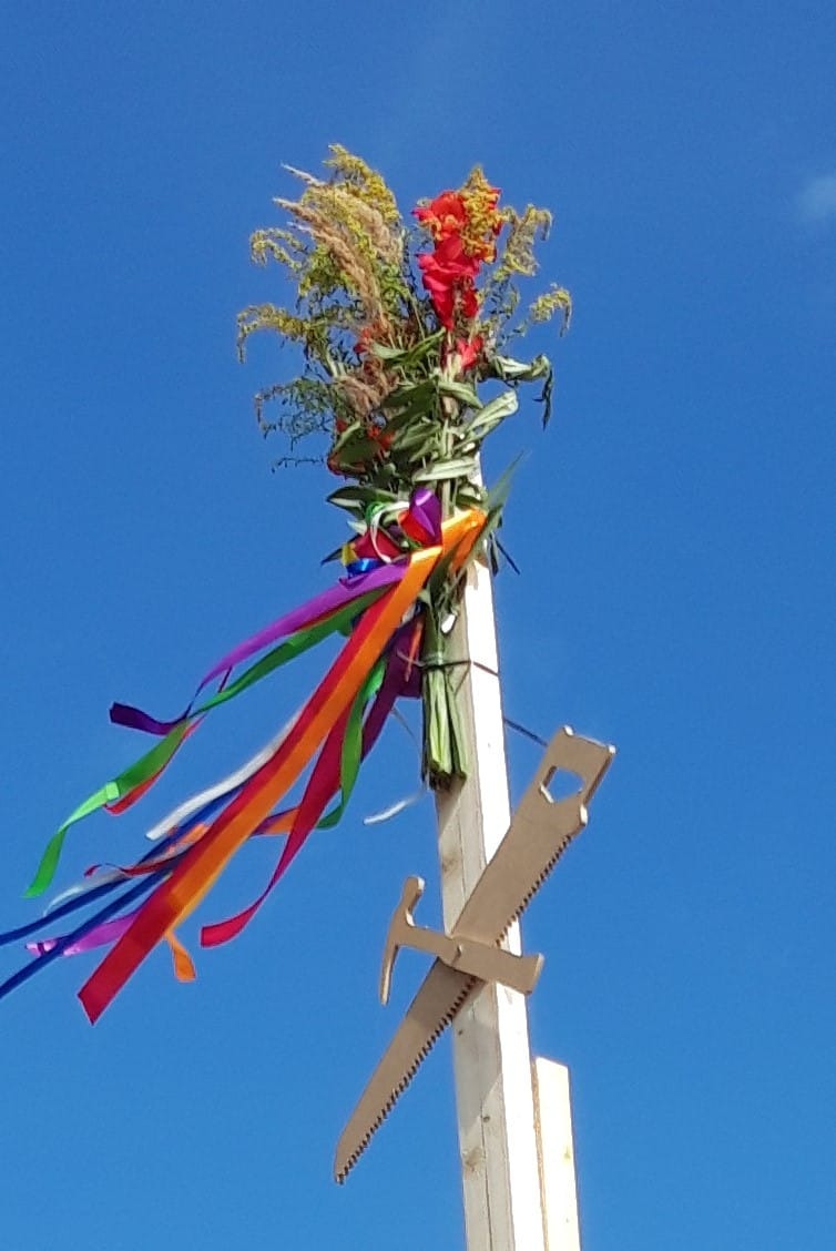 Majówka: drewniany słup z kwiatami, kolorowymi wstążkami i przytwierdzonymi narzędziami (piła, młotek) na tle błękitnego nieba. Nietypowa dekoracja budowlana.