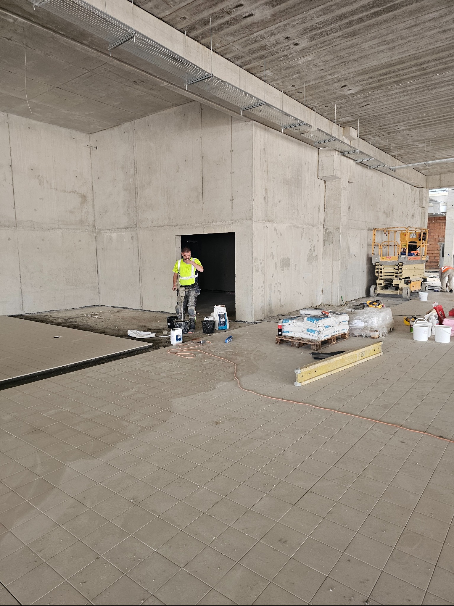 Układanie płytek podłogowych w surowym wnętrzu. Pracownik w odblaskowej kamizelce miesza zaprawę. Widoczne narzędzia, worki z zaprawą i podnośnik koszowy w tle.