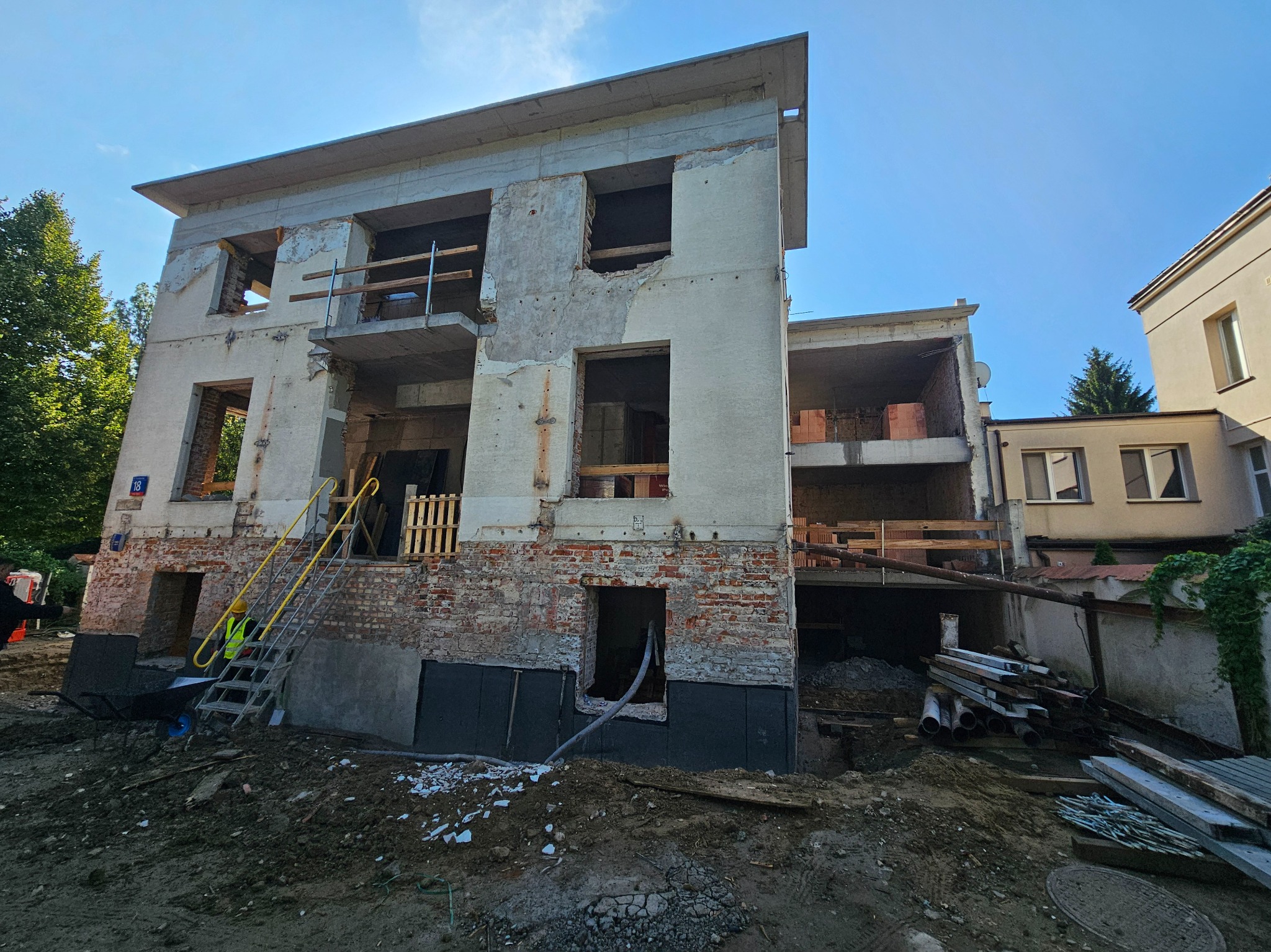 Budynek w trakcie rozbudowy z widoczną konstrukcją, ceglanym murem i schodami. Na pierwszym planie ziemia i materiały budowlane. Słoneczny dzień.