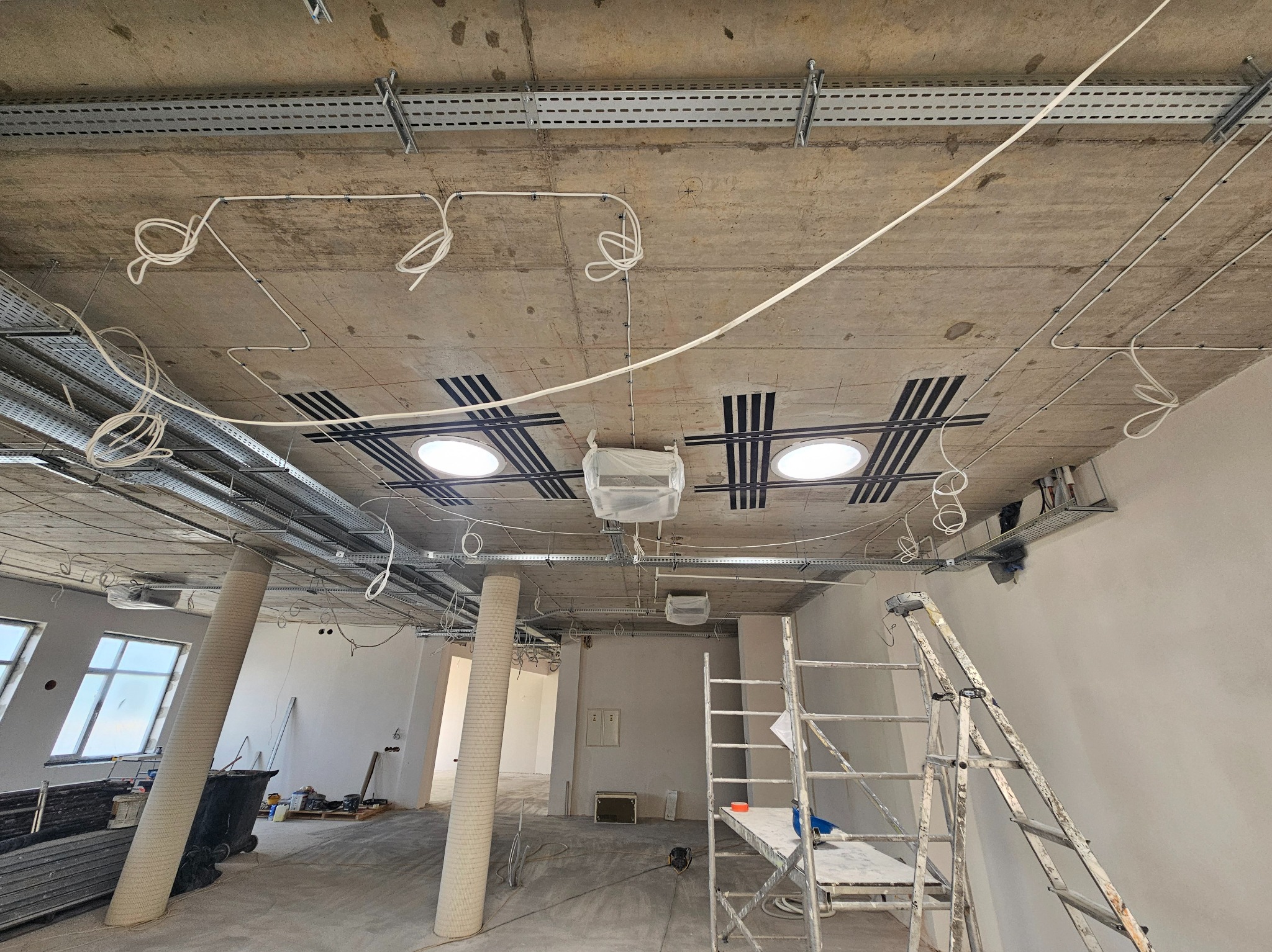 Instalacja elektryczna w biurze: kable w korytkach, oświetlenie sufitowe, drabina. Widok na sufit w trakcie prac wykończeniowych.