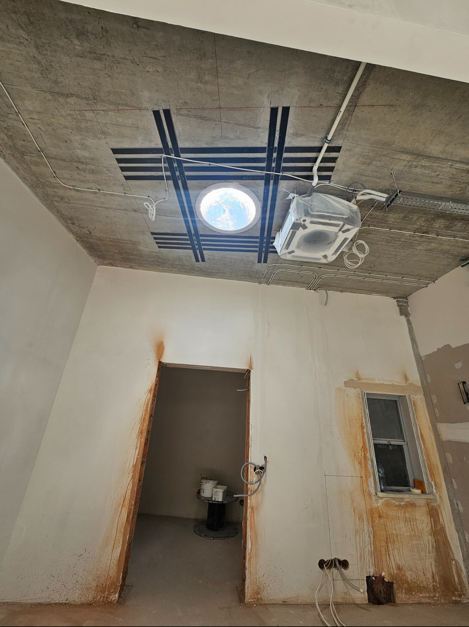 Wnętrze w trakcie remontu: surowy betonowy sufit ze wzmocnieniami, okrągłe okno dachowe, klimatyzator w folii, ściany w trakcie szpachlowania, widoczne instalacje.